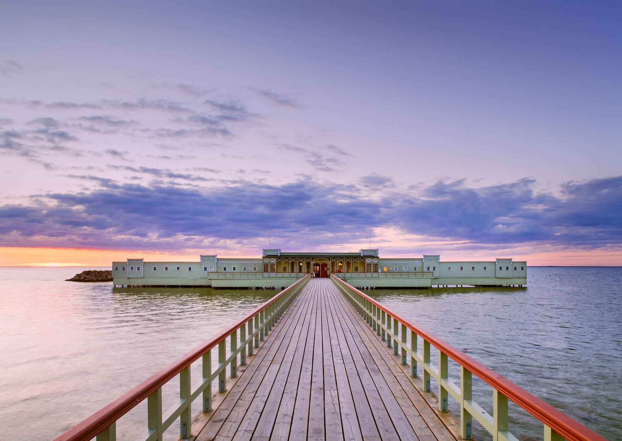 A long jetty in the water leading to a green open-air cold bath house.