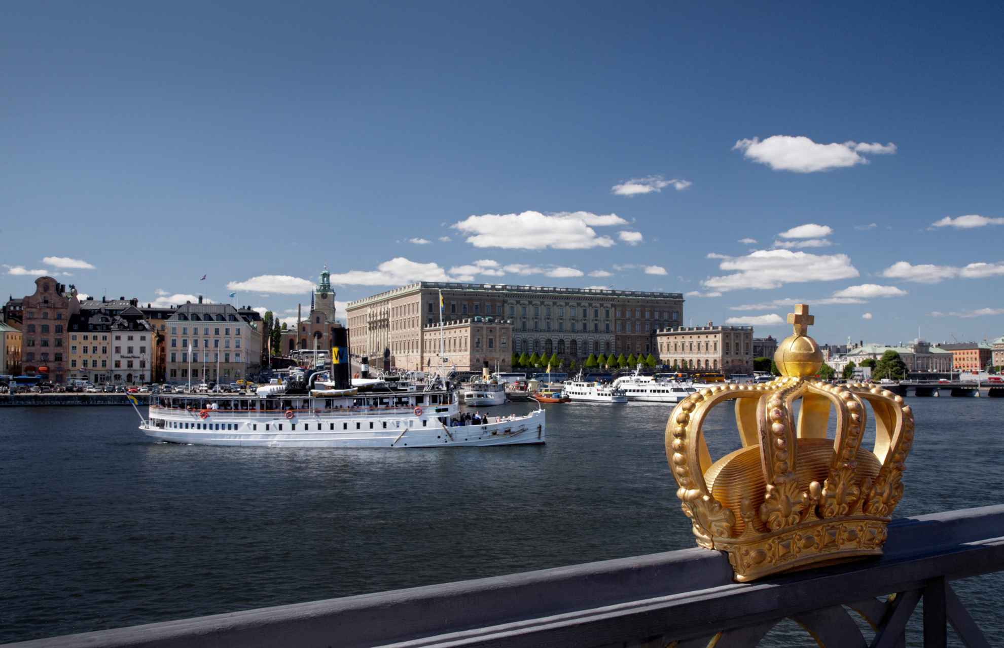 A golden crown fastened to the Skeppsholm Bridge with the Royal Castle and ferry boats in the background.