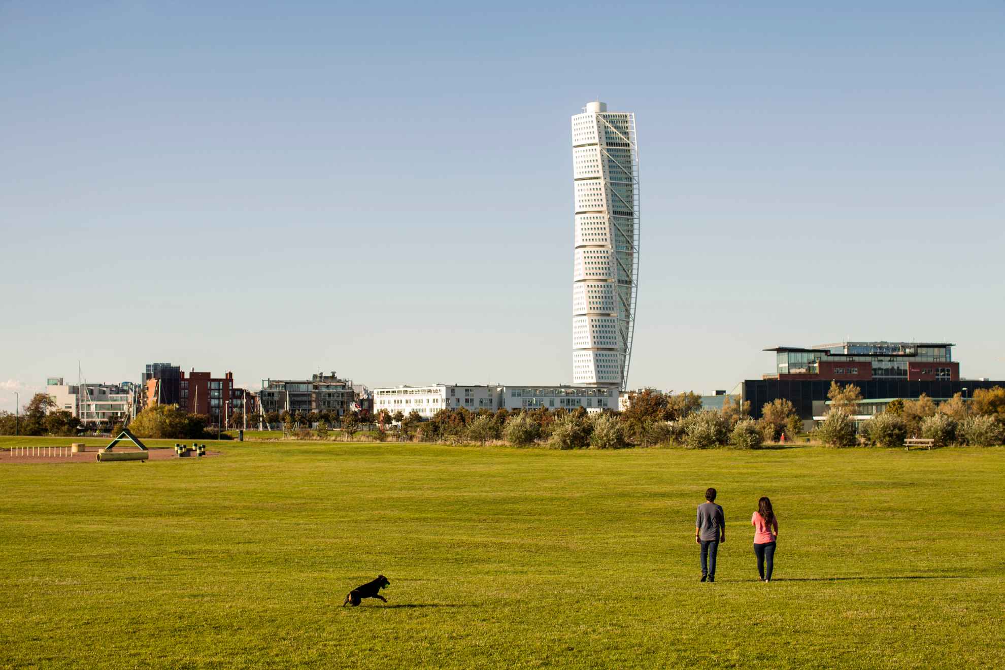 Two people are walking on a large field with their dog. The Turning Torso can be seen in the background.