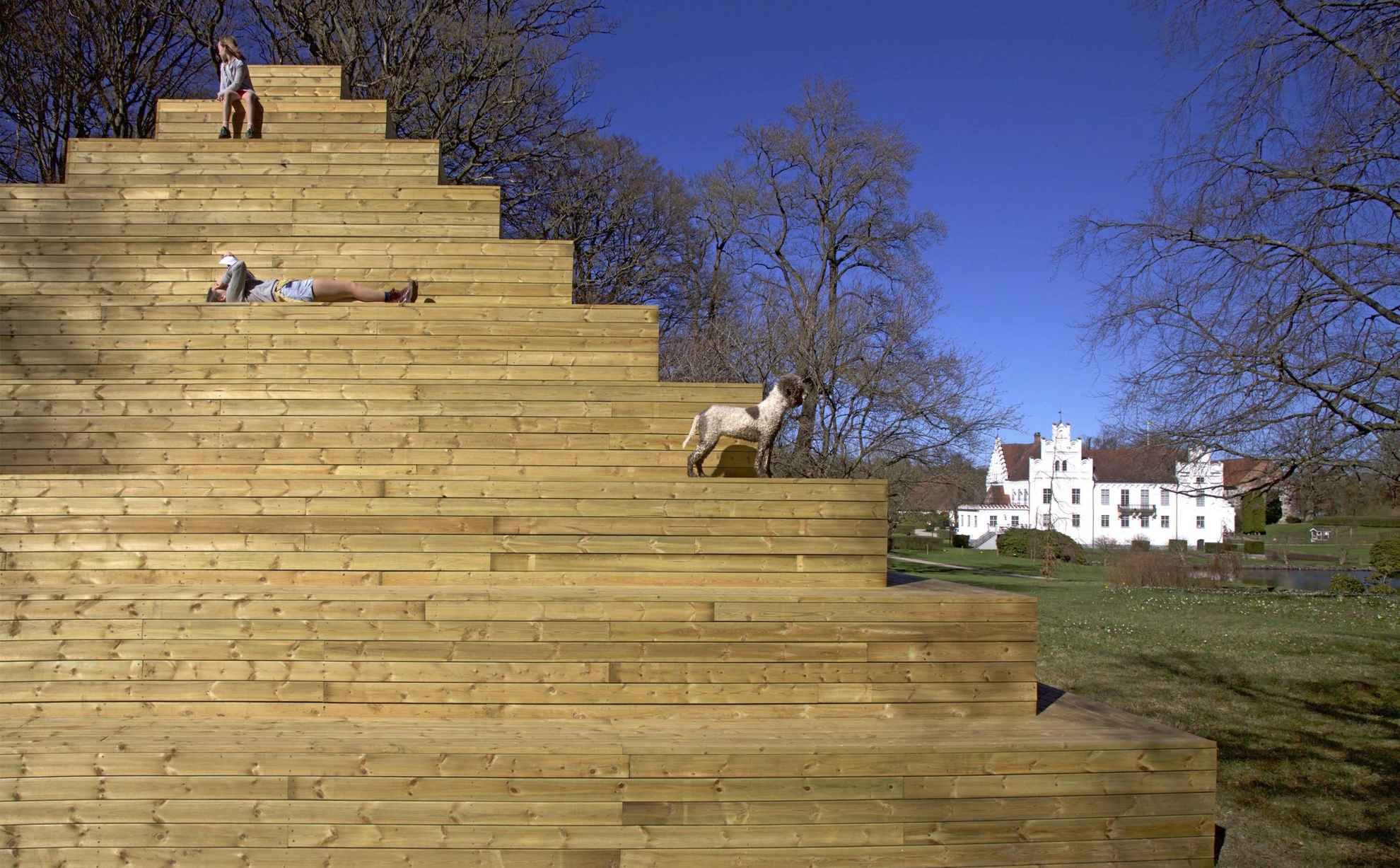 A sculpture named  Pyramide, made in wood as a place “where people would be able to hang out”. A dog standing on the pyramid, a person is sitting on the top and another one is lying in the middle. A large white house in the background.