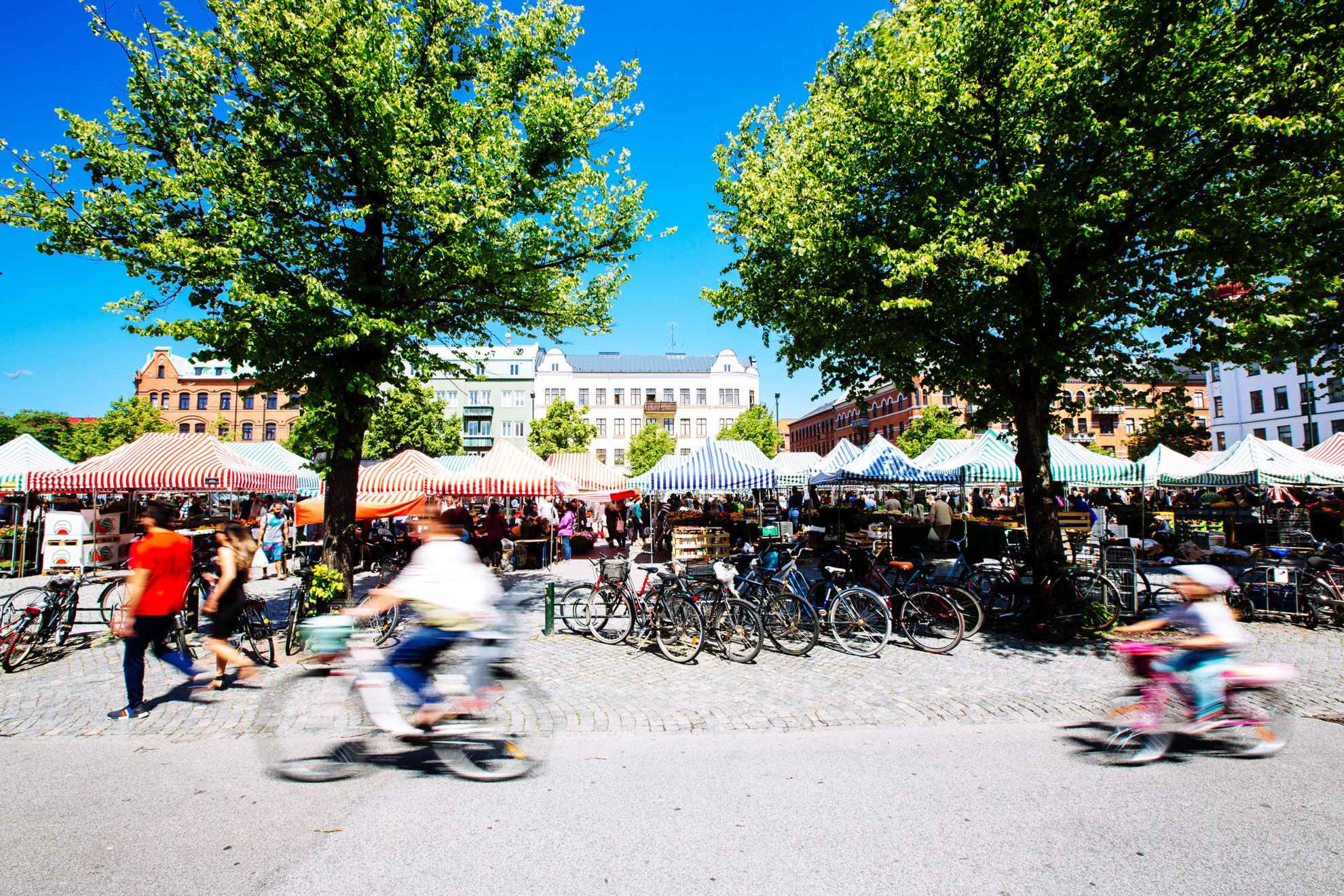 A square full of stalls with striped roofs. Bikes are parked along a road and you see two blurry cyclists  passing by.