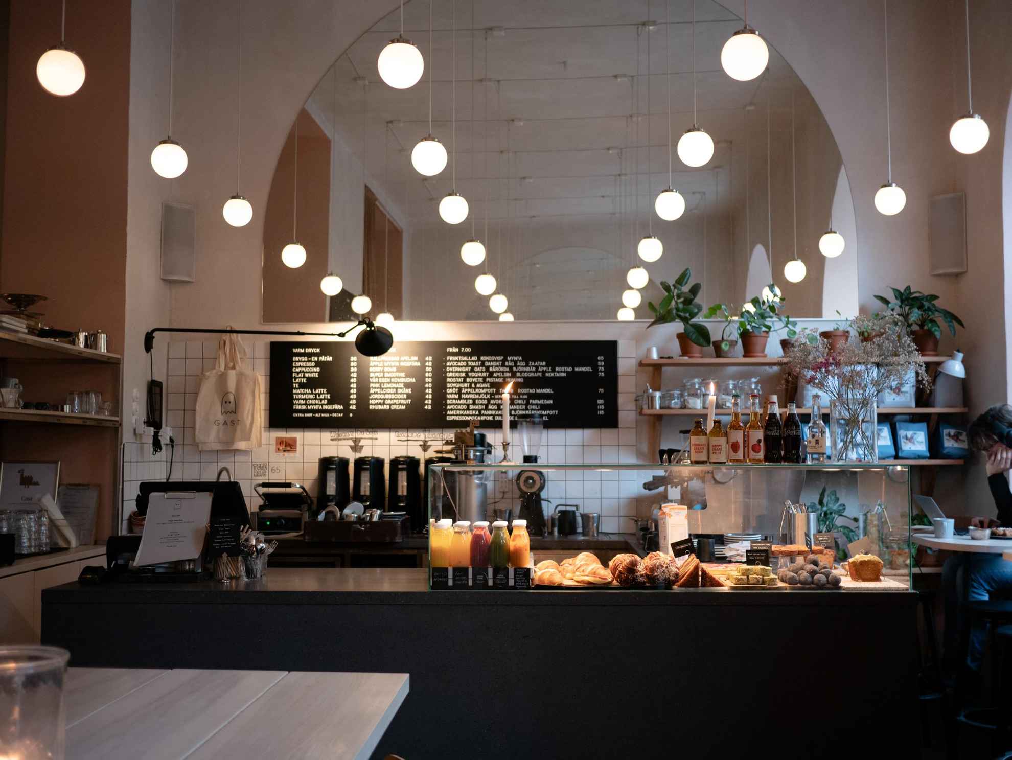 A serving counter with buns, bread, pastries and various soft drinks in a cafe. Behind the counter a coffee machine. In the ceiling hangs several small globe lights.