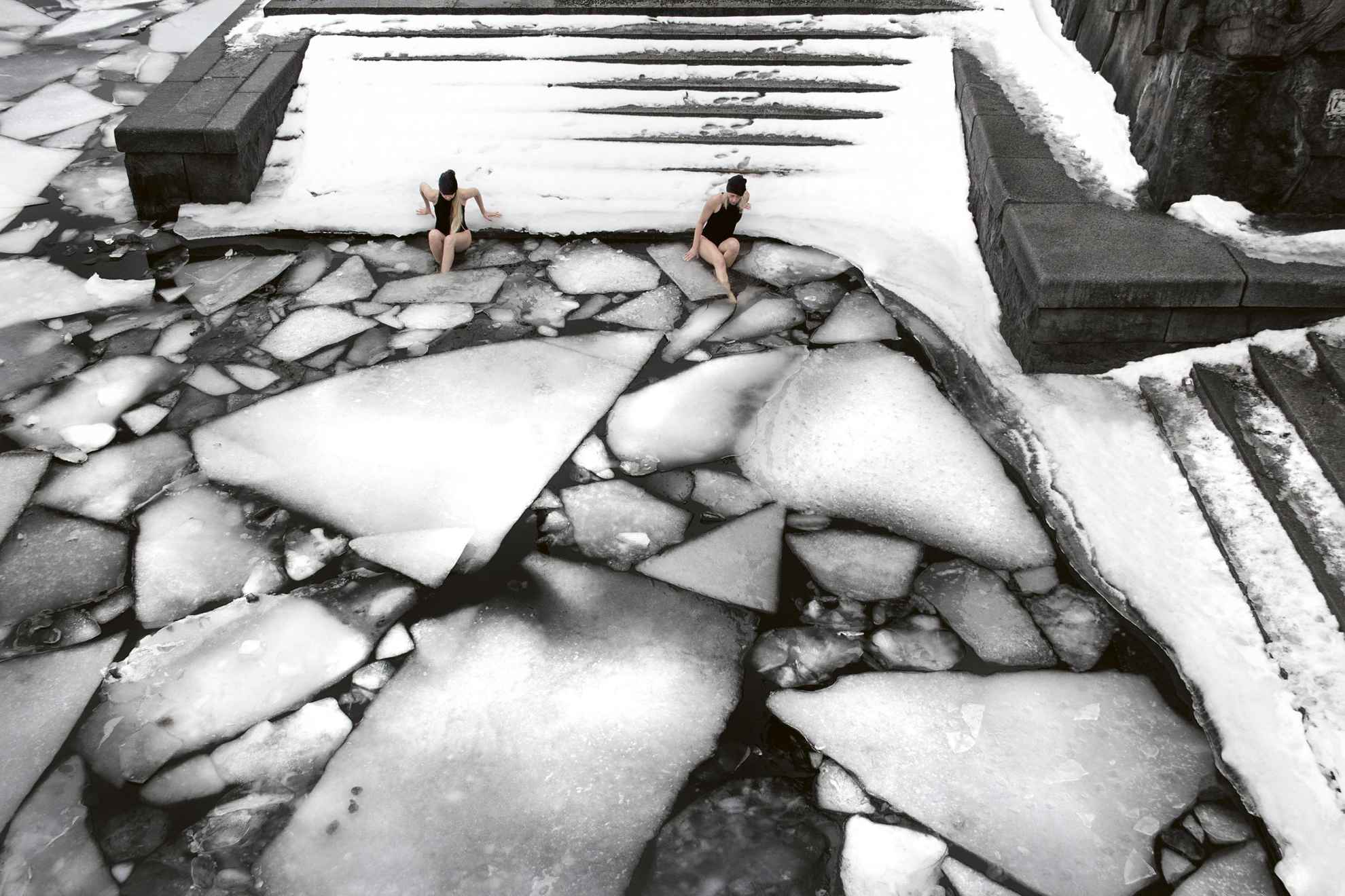 Two women on their way to take a winter bath from a jetty with stairs down to the shore. There are ice floes in the water.