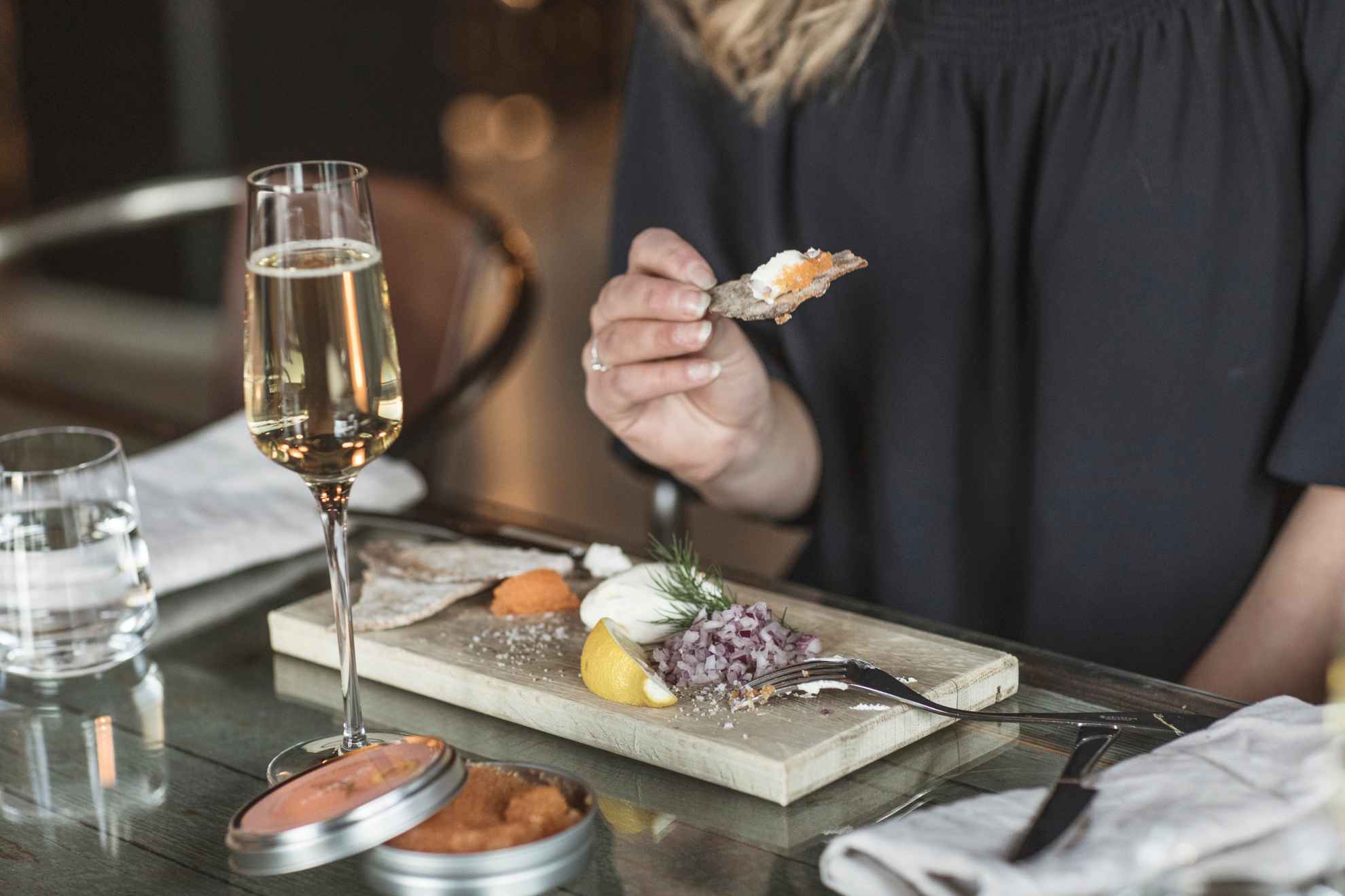 Close-up of a woman eating caviar, red onion, sour cream, lemon and crispbread from a wooden tray and drinking sparkling wine.