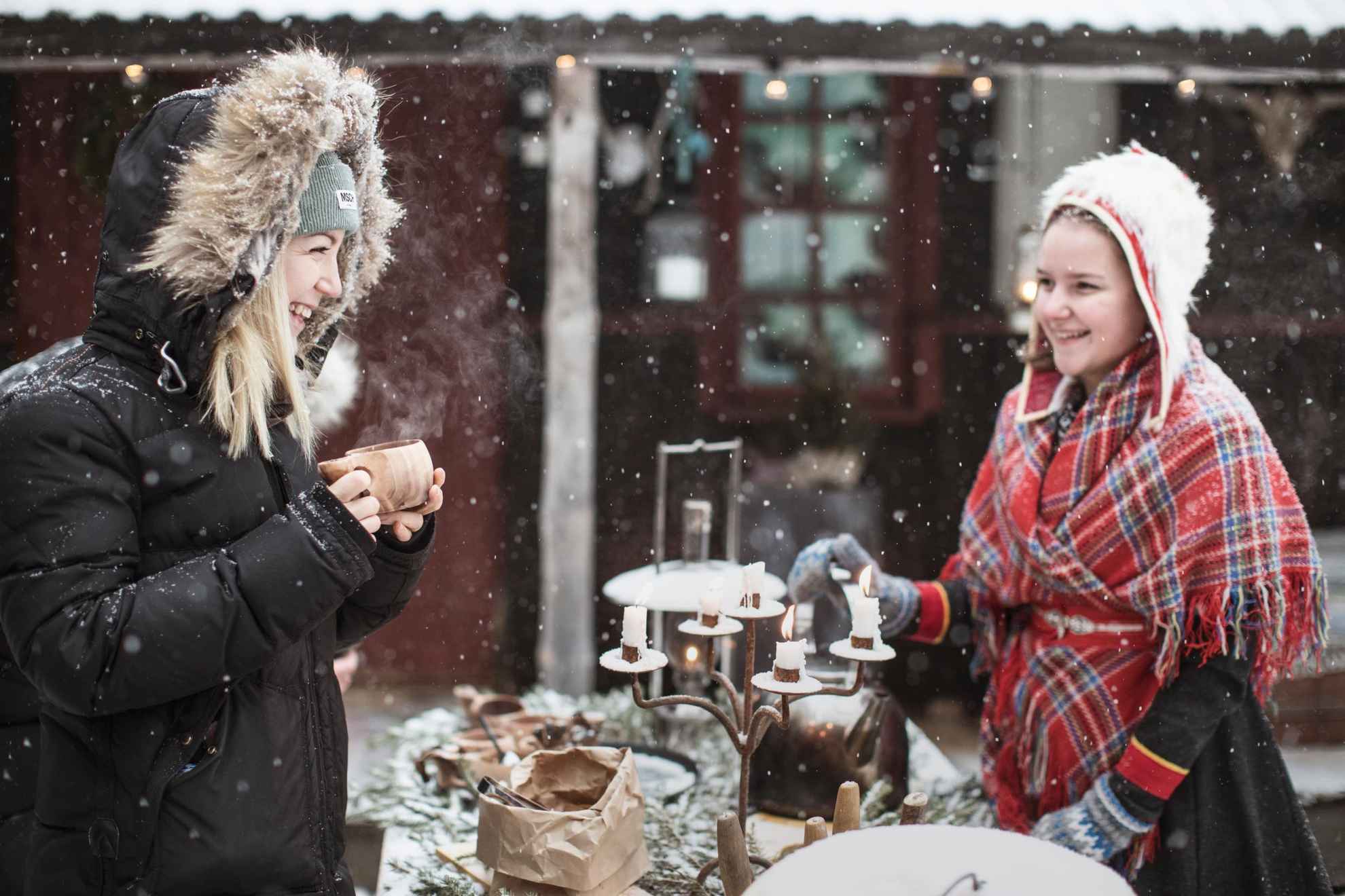 Two women  stands at a table having an outdoor fika in the snow. There are an old candelabra with lit candles on the table. One of the girls are wearing a Sámi costume. It’s snowing.