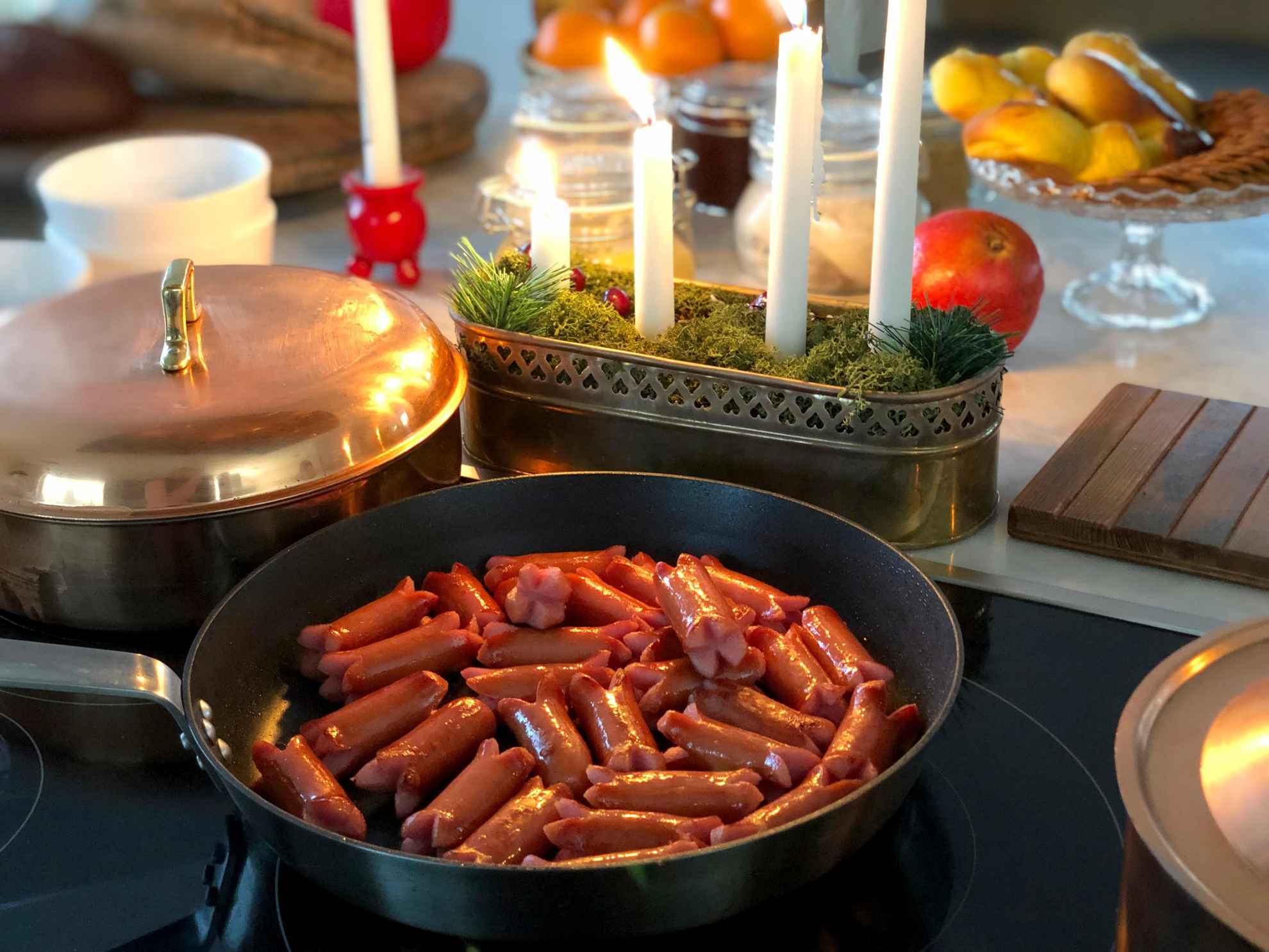 Close up of a frying pan with prince sausages next to an advent candlestick with lit candles and a copper pot. Fruits in the background.