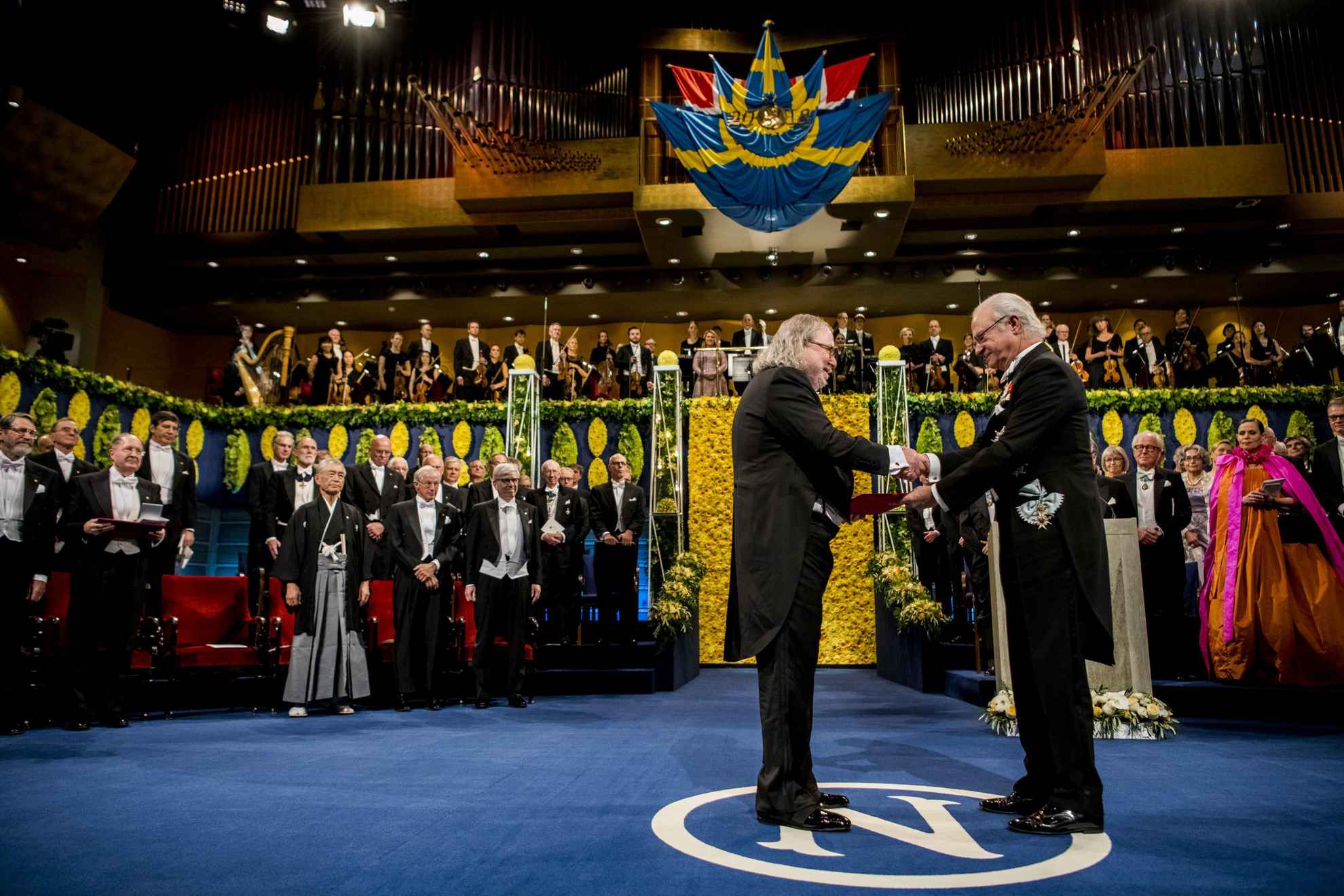 A male laureate is standing on a stage and receives his prize from the Swedish King Carl XVI Gustaf.