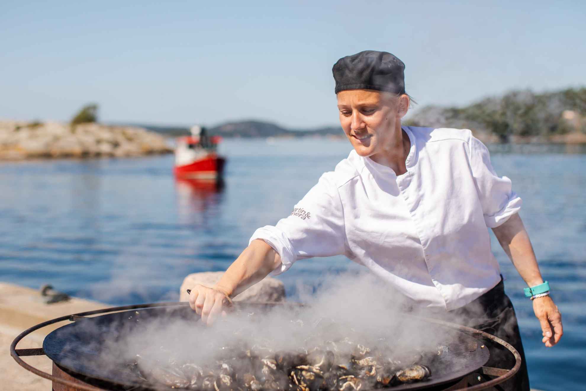 A chef is standing outside by the sea cooking mussels in a large pan.