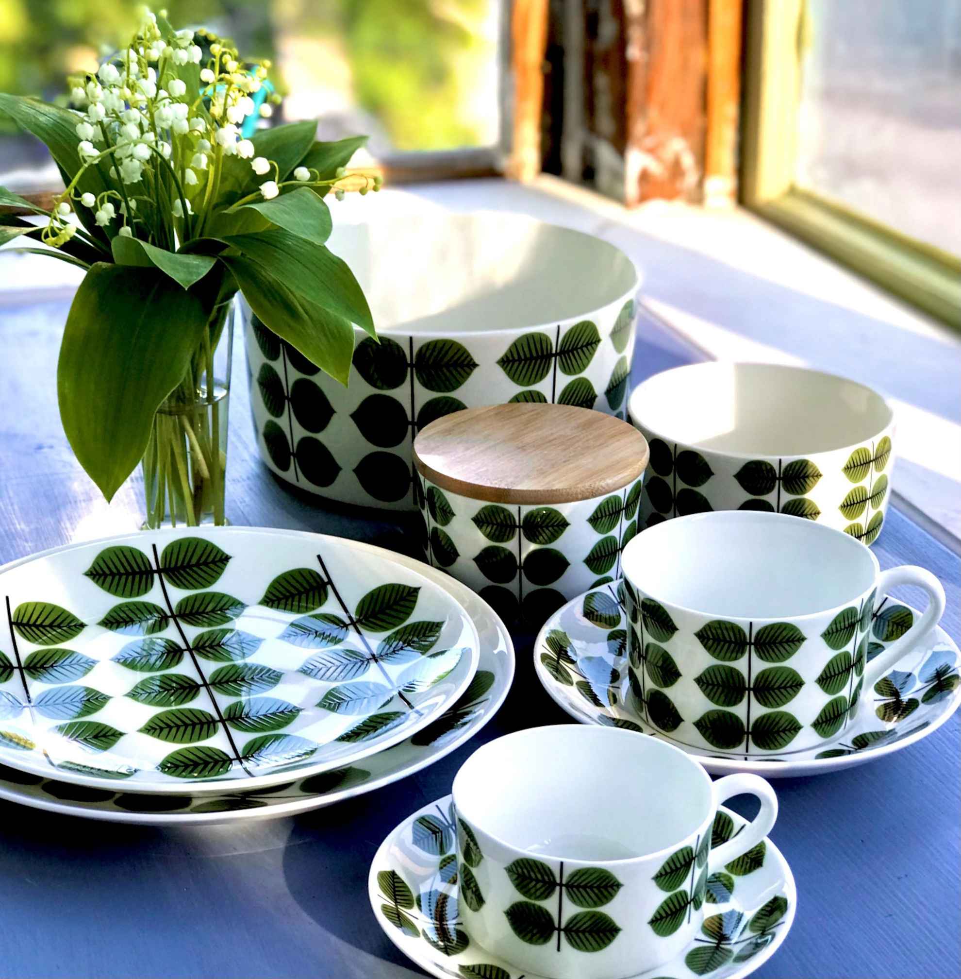 Close-up on a set with porcelain products painted with green leaves and a vase with lilies of the valley on a table.