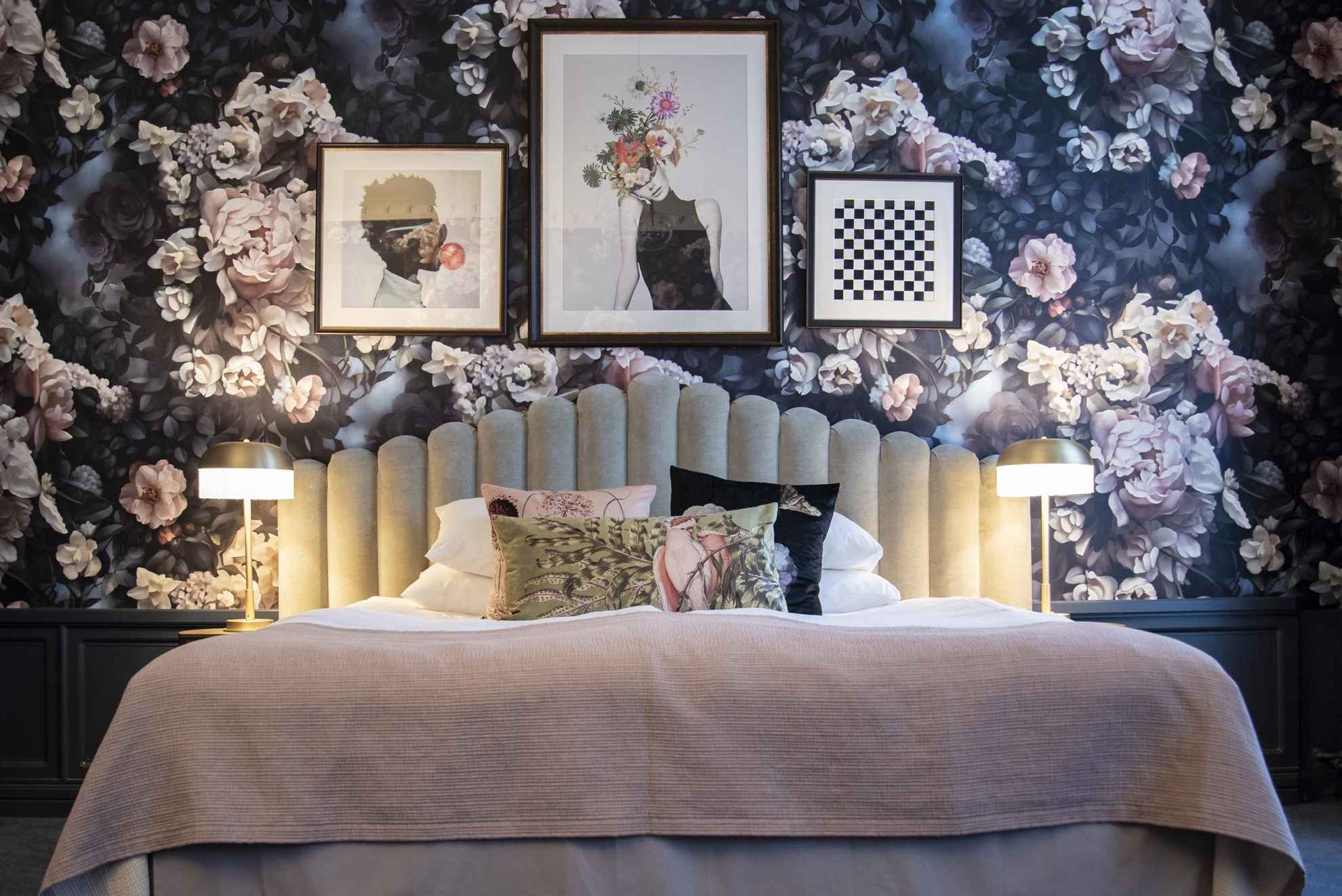 Inside a room at a hotel. A bed in front of a wall with a wallpaper with large pink and white roses. There are three pictures on the wall and three cushions in different pattern on the bed.