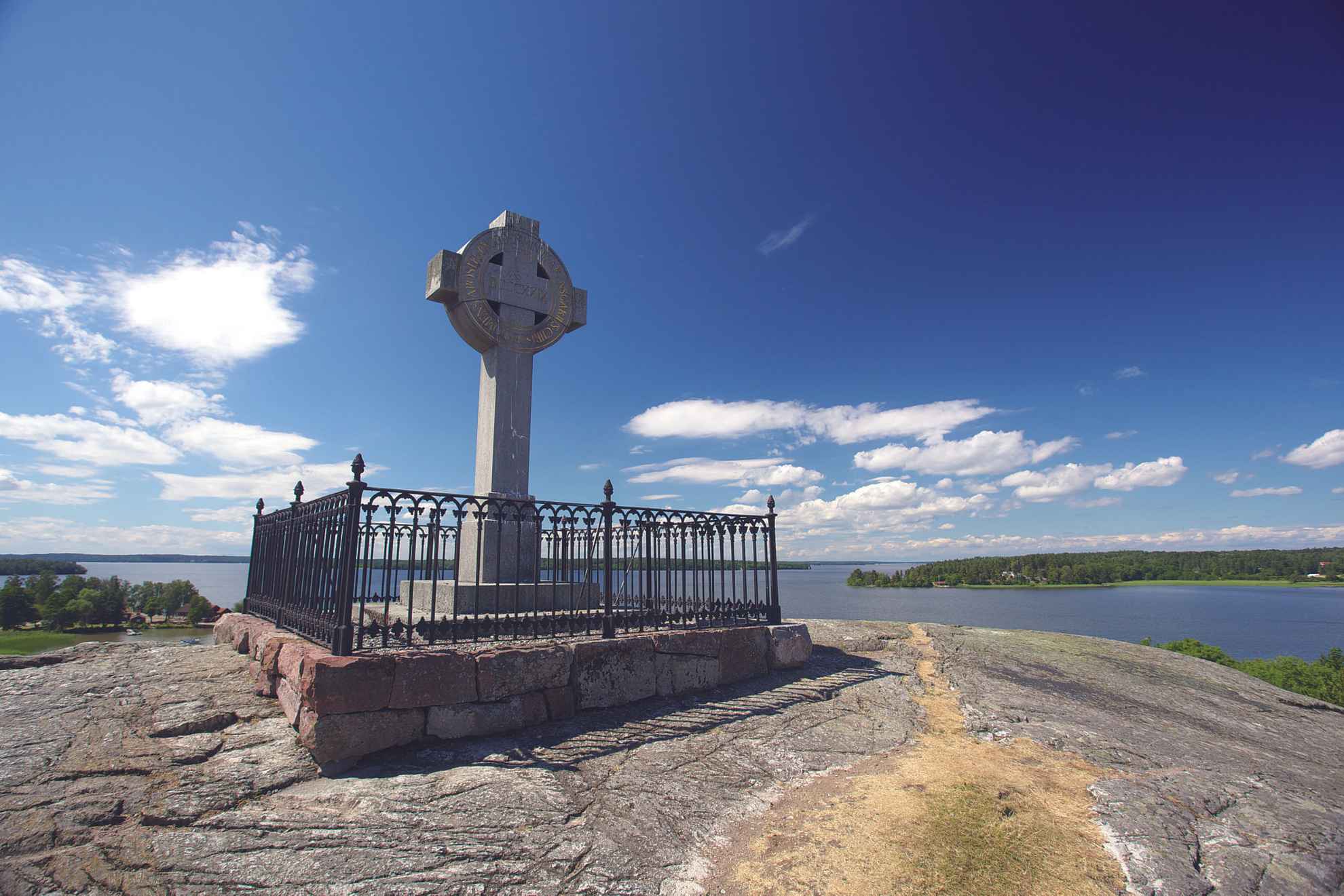 The Ansgar Cross stands on top of a bare cliff. You can se the lake Mälaren, islets and a blue sky in the background.