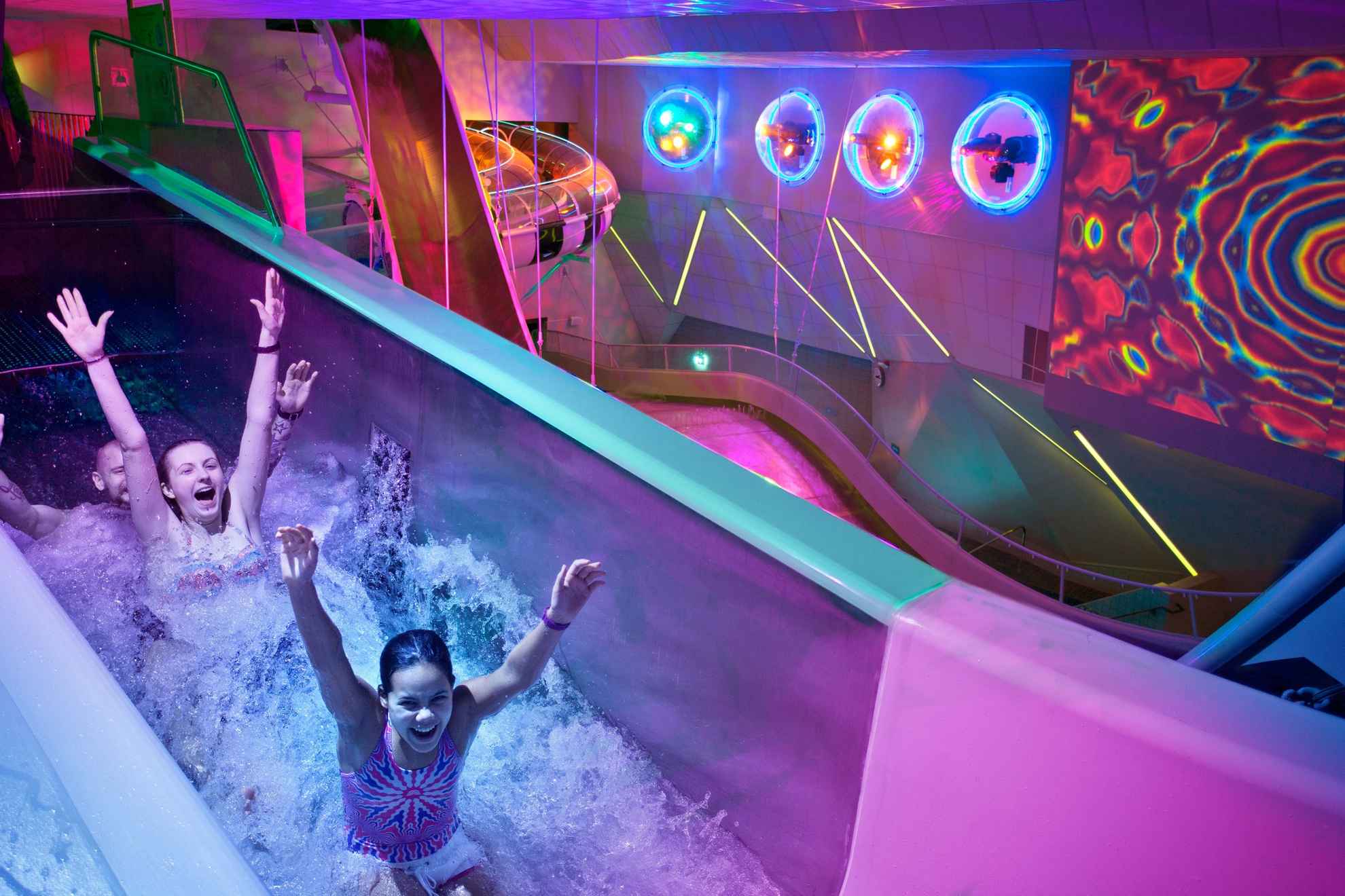 Three people smile and hold up their arms as they go down a water slide. The area is mostly in purple and neon lights.