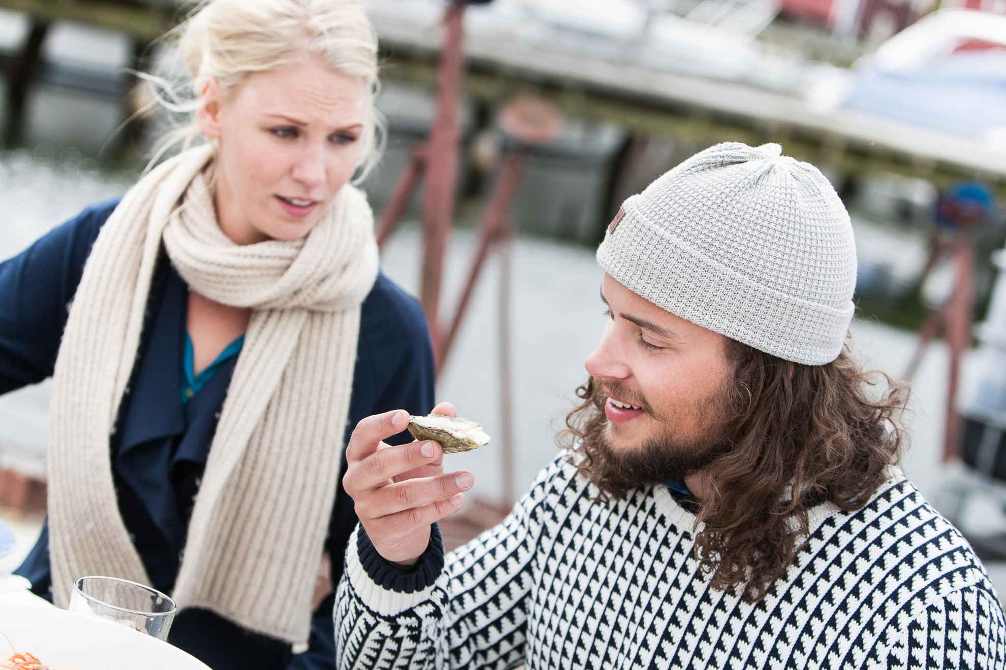 A man and a woman sits at a table outdoor. The man have an oyster in his hand.