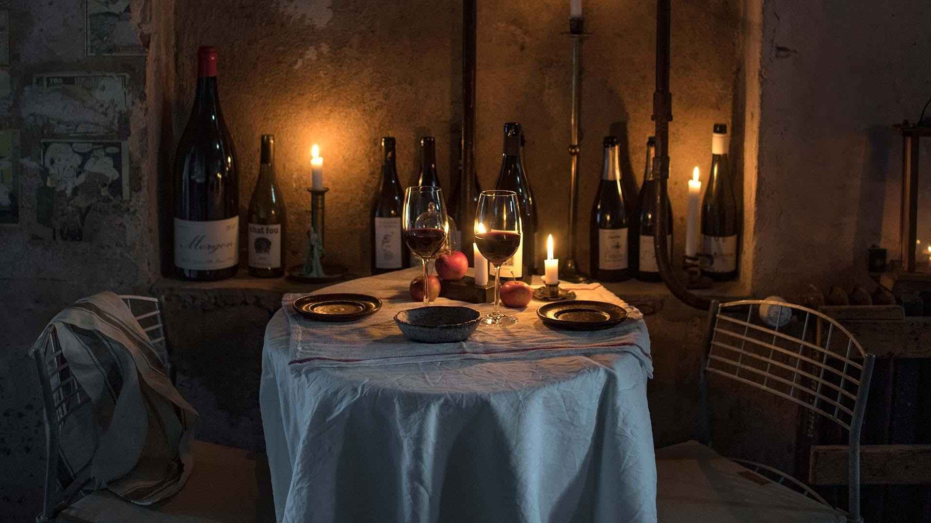 A table for two set with a white tablecloth, plates and two glasses filled with red wine. The wall is decorated with empty wine bottles and lit candles.