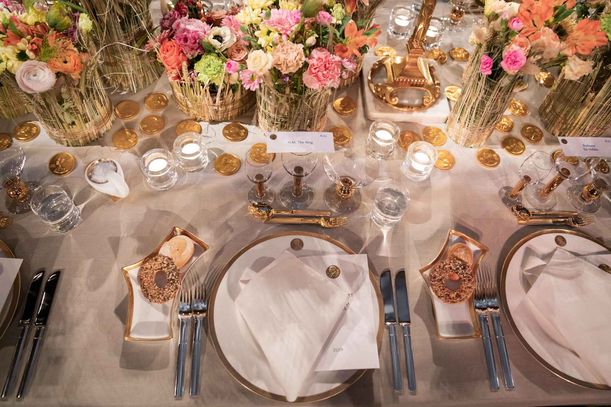 A tableware in white with golden details is set on a table with a white tablecloth. In the middle of the table are colorful flowers in vases.