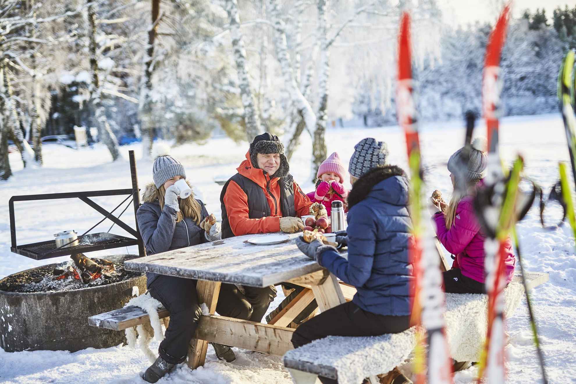 A family of five sitting outside at a table, eating cinnamon buns and drinking coffee. There are skis in the foreground of the picture.