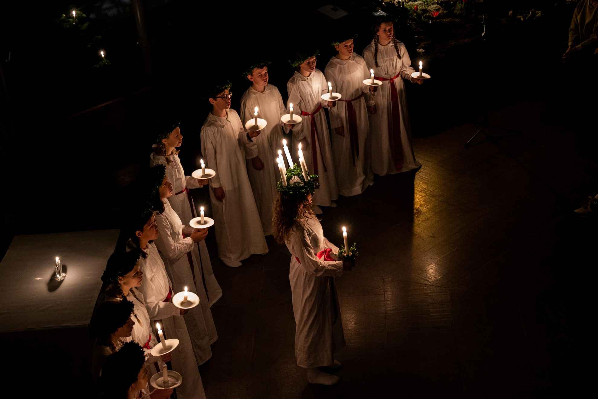 A Lucia with candles in her hair seen from the side and behind her several handmaidens all carrying a candle in the hand and dressed in white gowns.