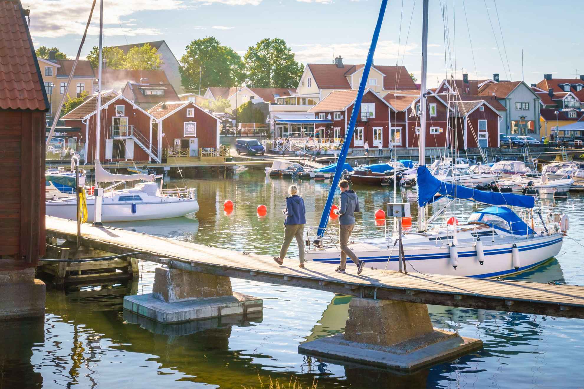 A couple walks on a jetty in a marina in a small wooden city. There are red cottages along the waterside.