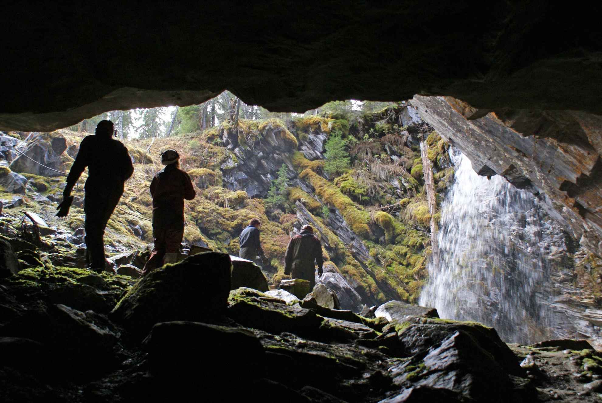 A view from inside a cave out into a glade with four people walking towards a waterfall.