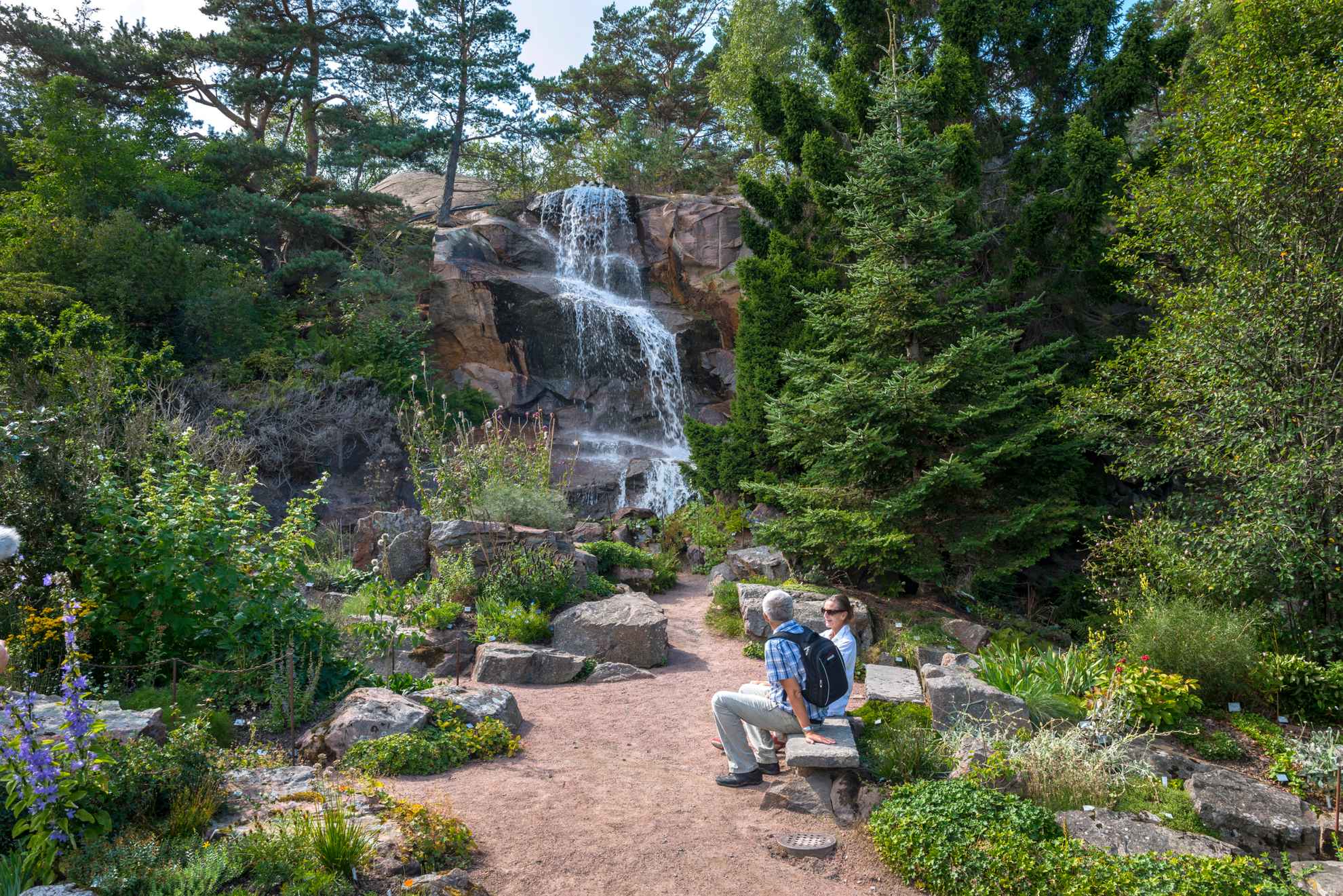 A couple sits on a park bench overlooking a small artificial waterfall in Gothenburg Botanical Garden.