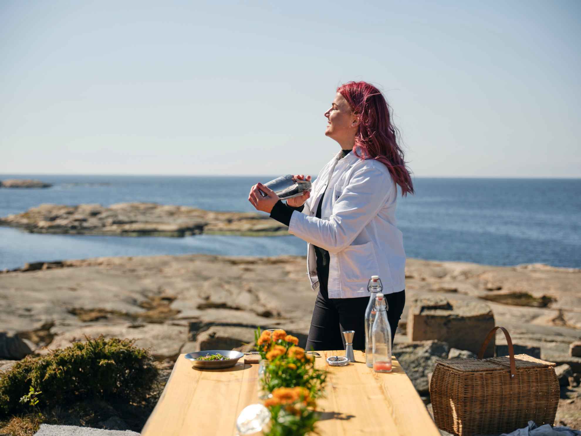A female bartender is mixing a drink next to a wooden table on the cliffs at the coast. The table is decorated with orange flowers and there are two bottles and a plate on the table.