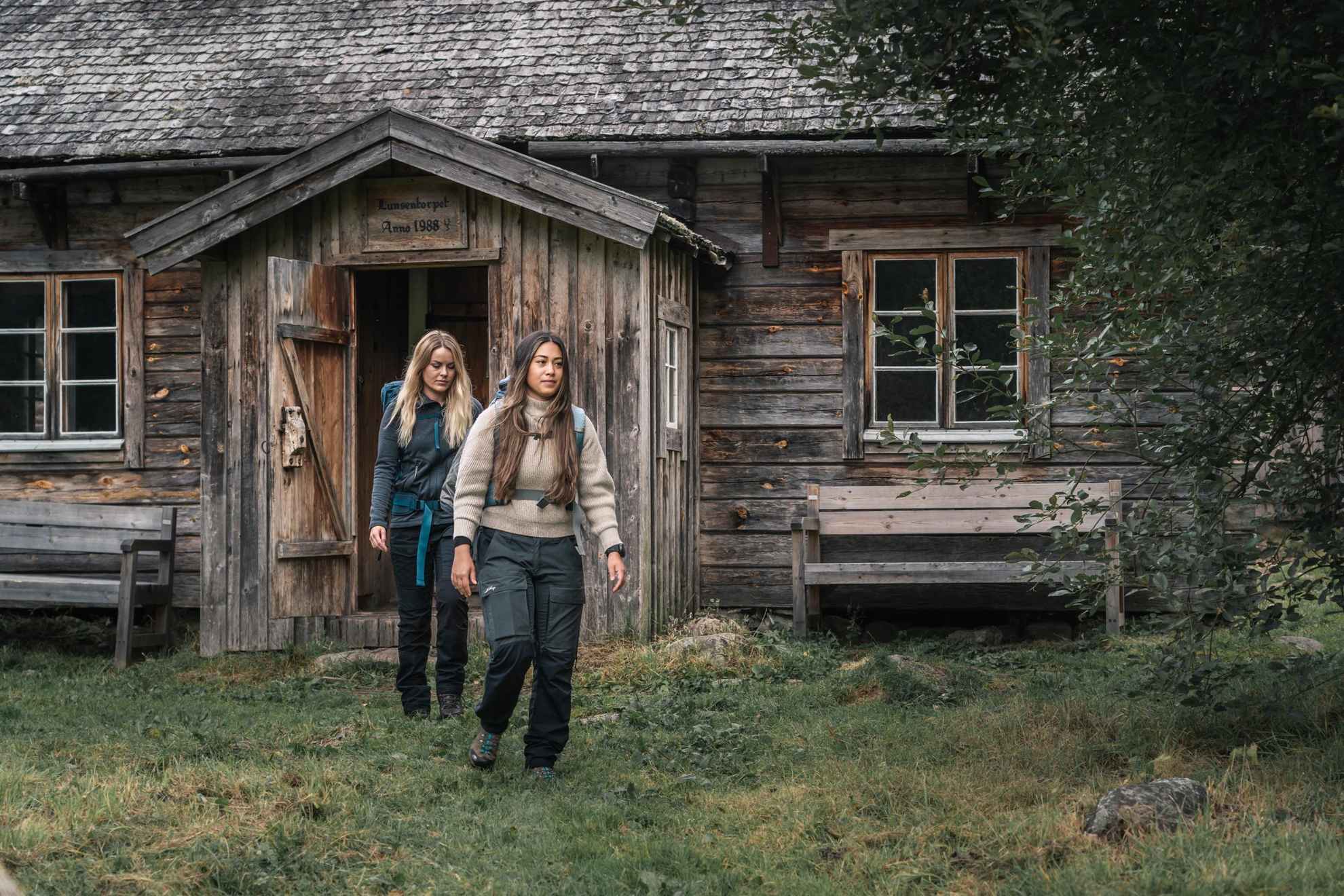 Two hiking women are leaving a wooden cottage in the forest.