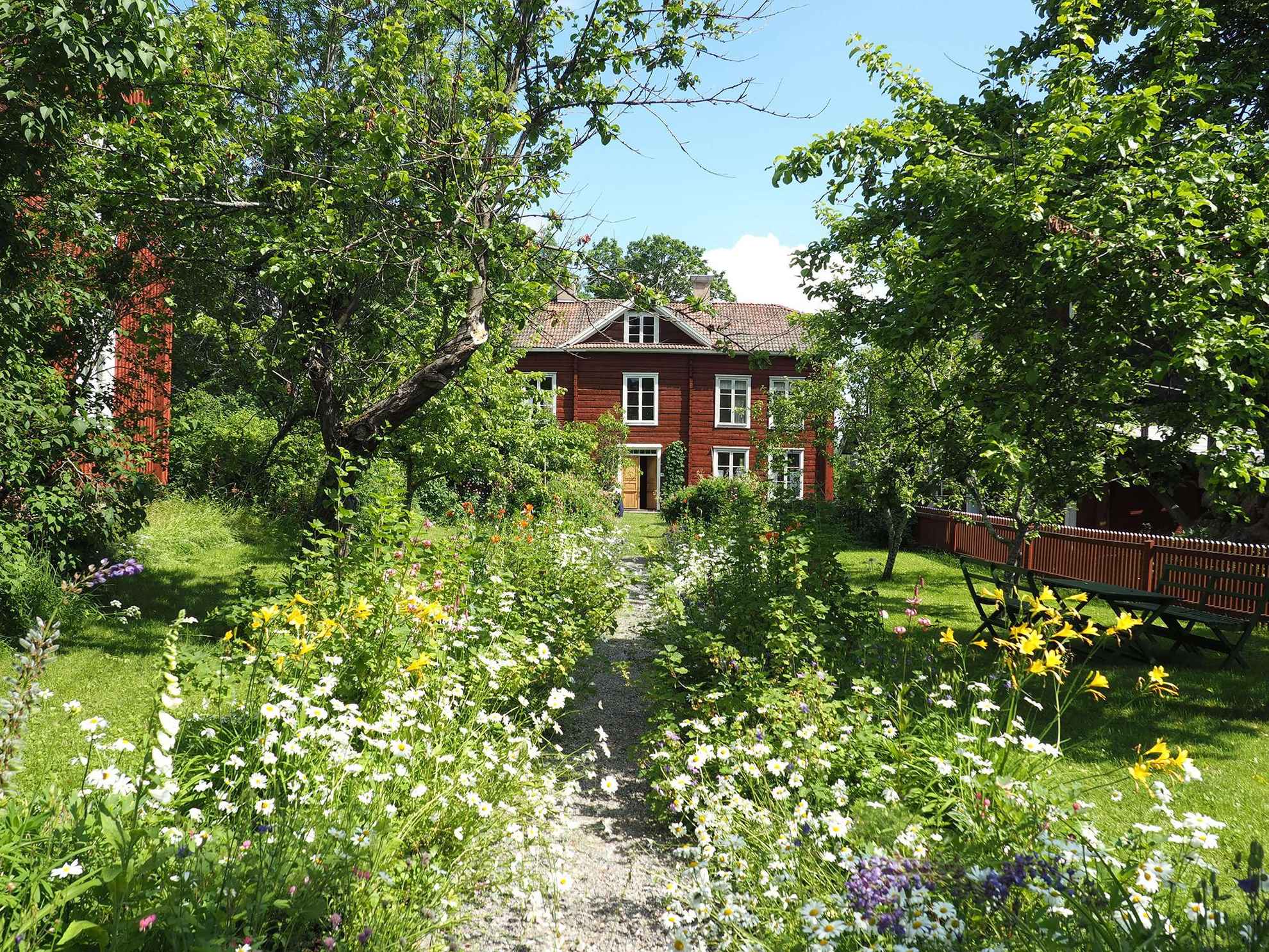 A gravel road with blooming flowers on each side leads up to a red wooden house. There is outdoor furniture and trees on the green lawn.