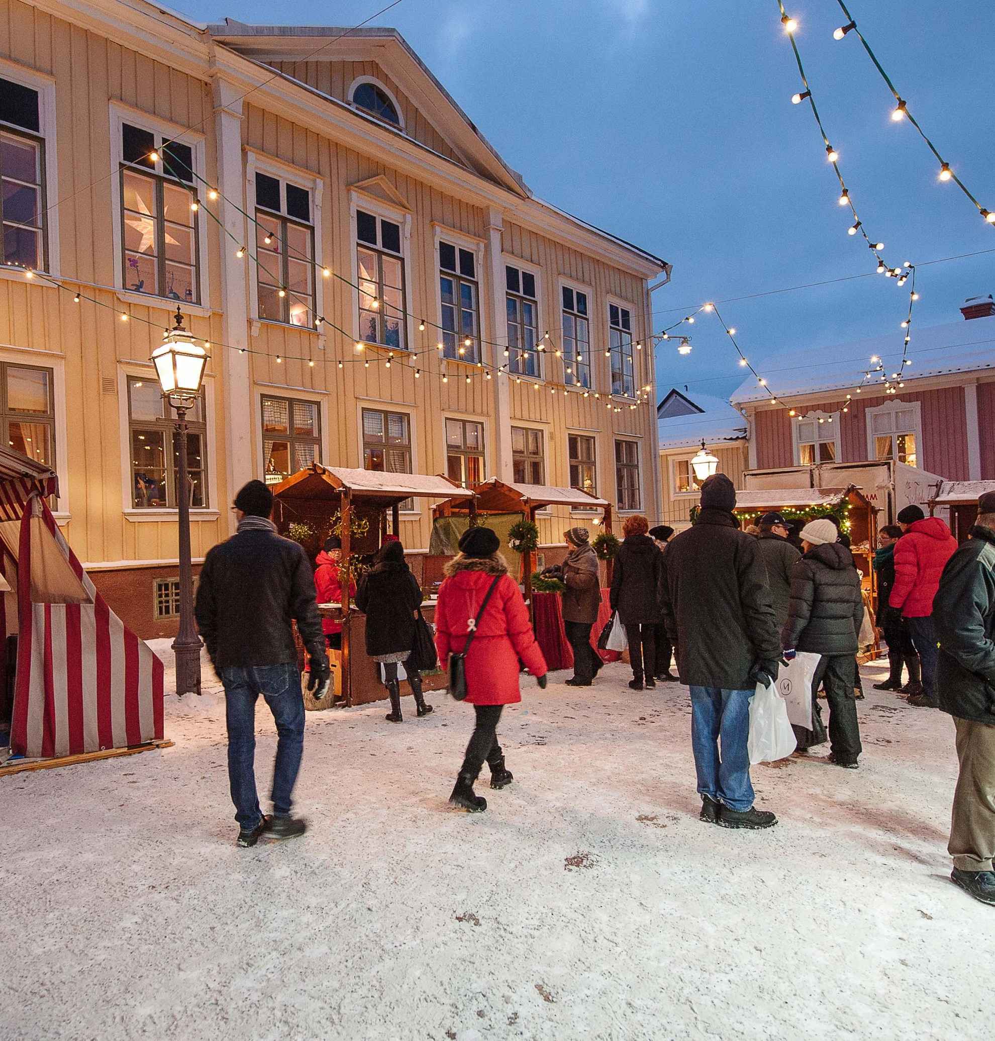 People are walking around the stables in front of a yellow wooden building at Eksjö Christmas market. It's getting dark, lights decorations have been lit and there's snow on the ground.