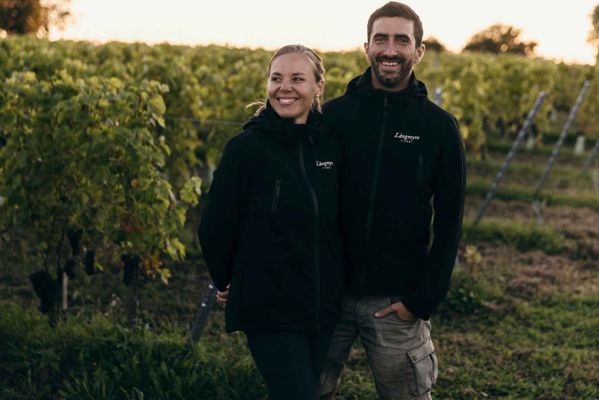 Emma Serner and Andrea Guerra standing in front of some green grapevines at their vineyard, Långmyre.