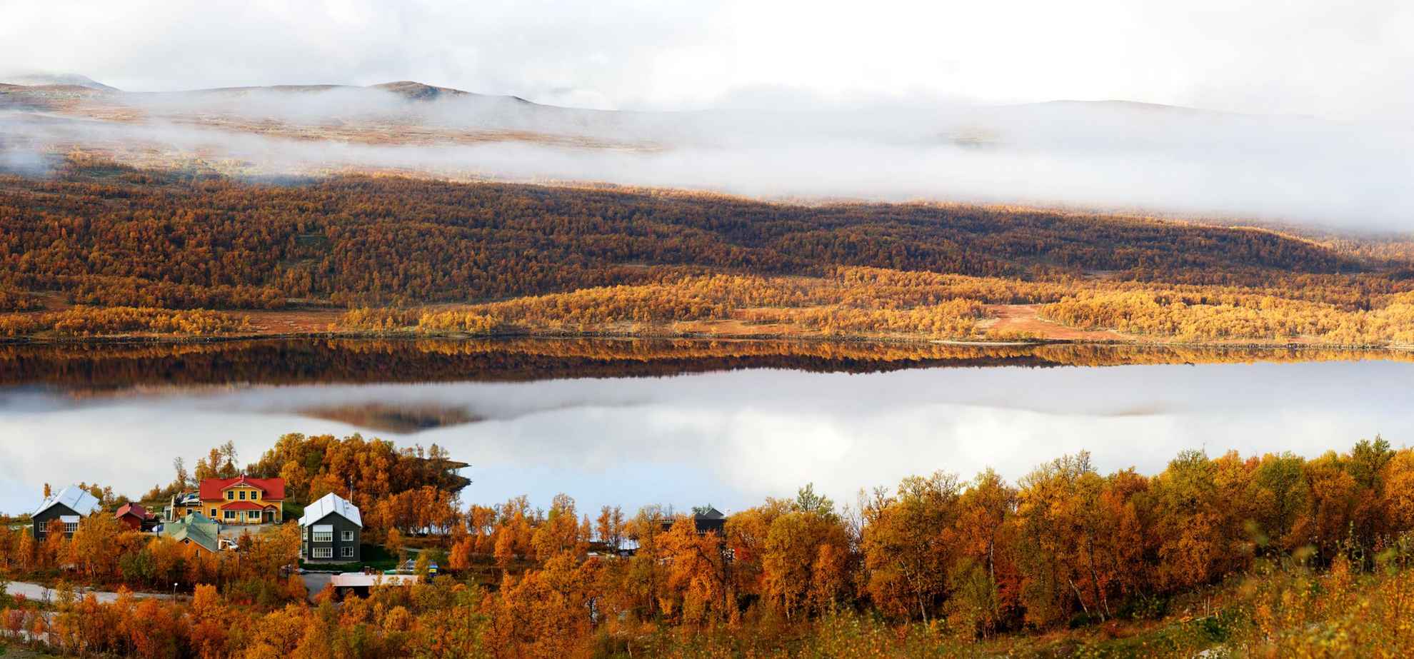 A beautiful autumn view of a hotel by a lake in the mountains. The trees are orange and there is a fog over the mountain on the other side of the lake.