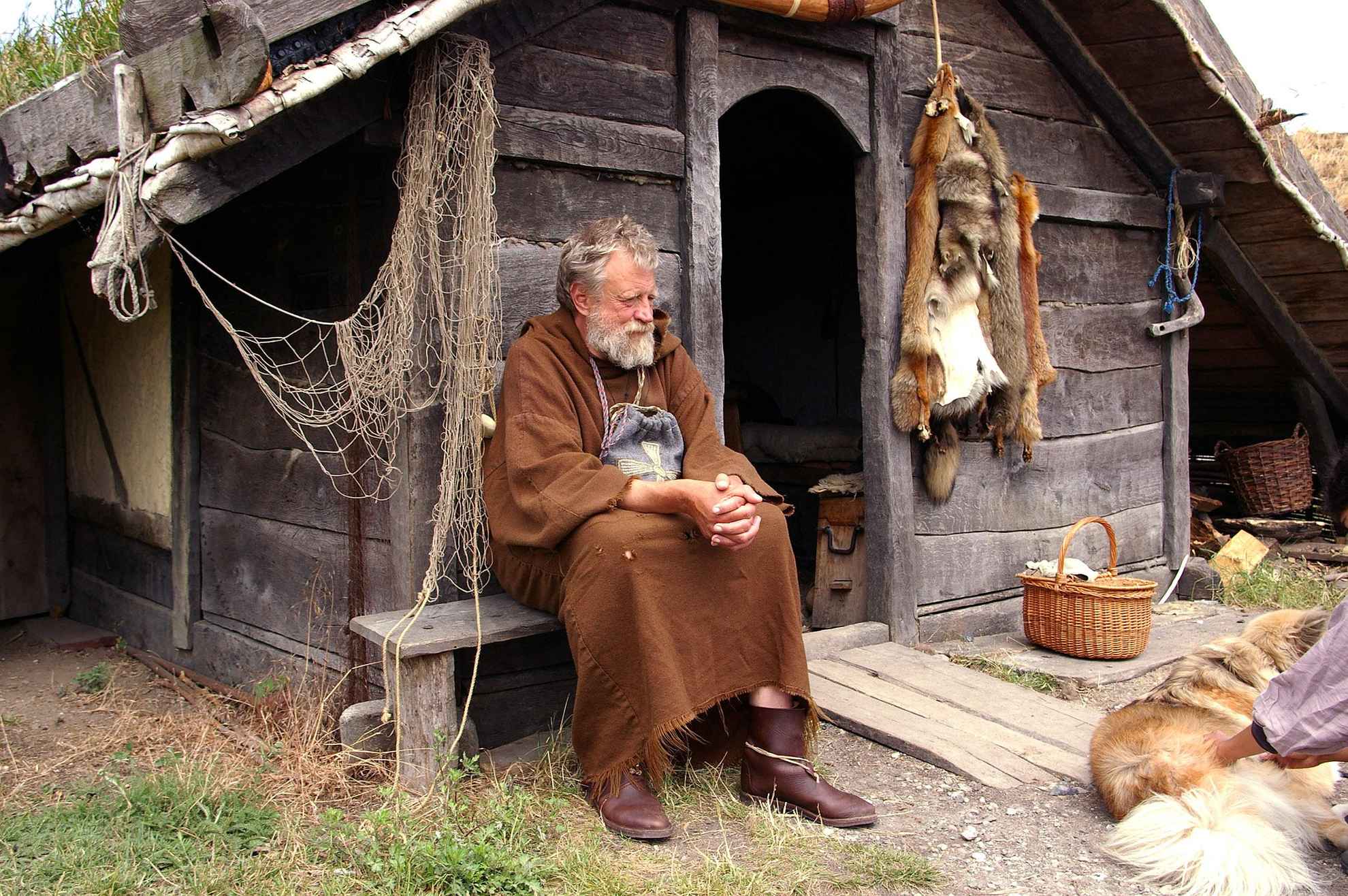 An old man dressed as a viking sits in front of an old wooden house.