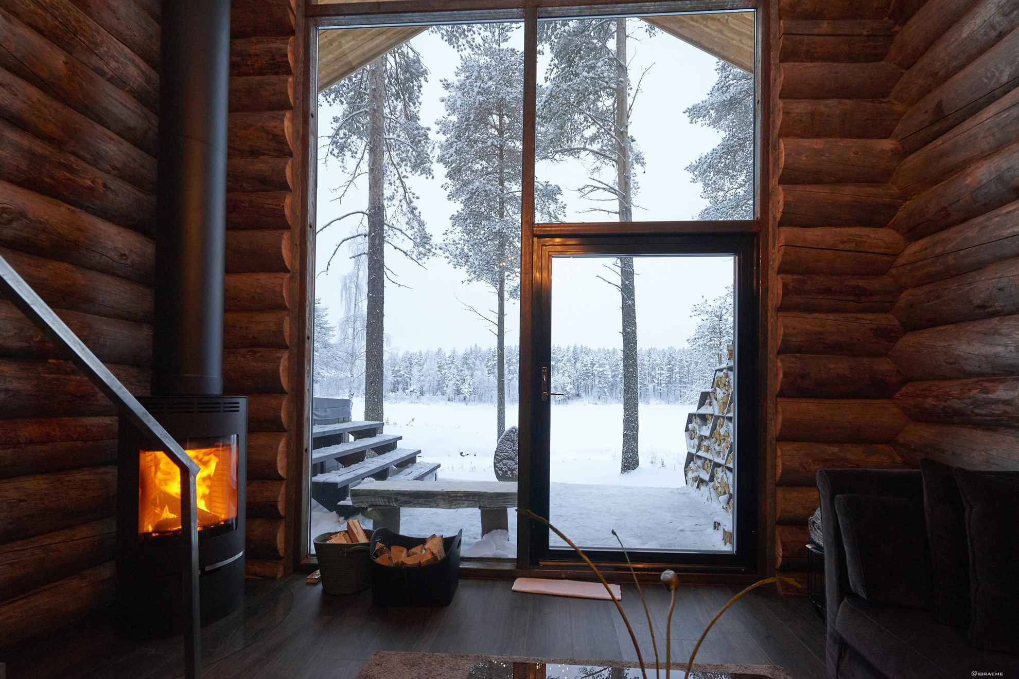A timber log cabin with a fireplace and large windows. It is winter outside.