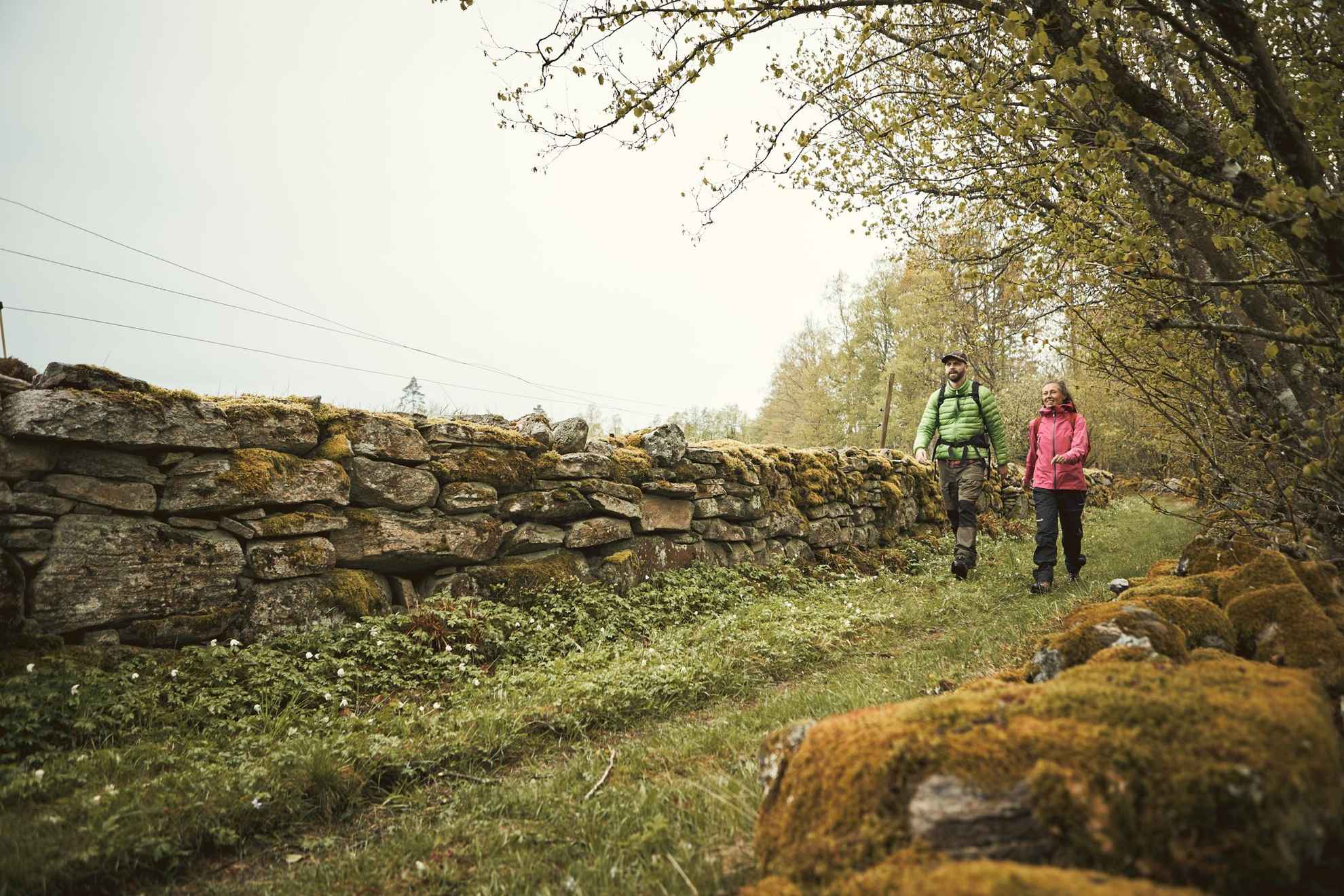 A man and a woman is hiking on a trail next to a stone wall and a forest on the other side.