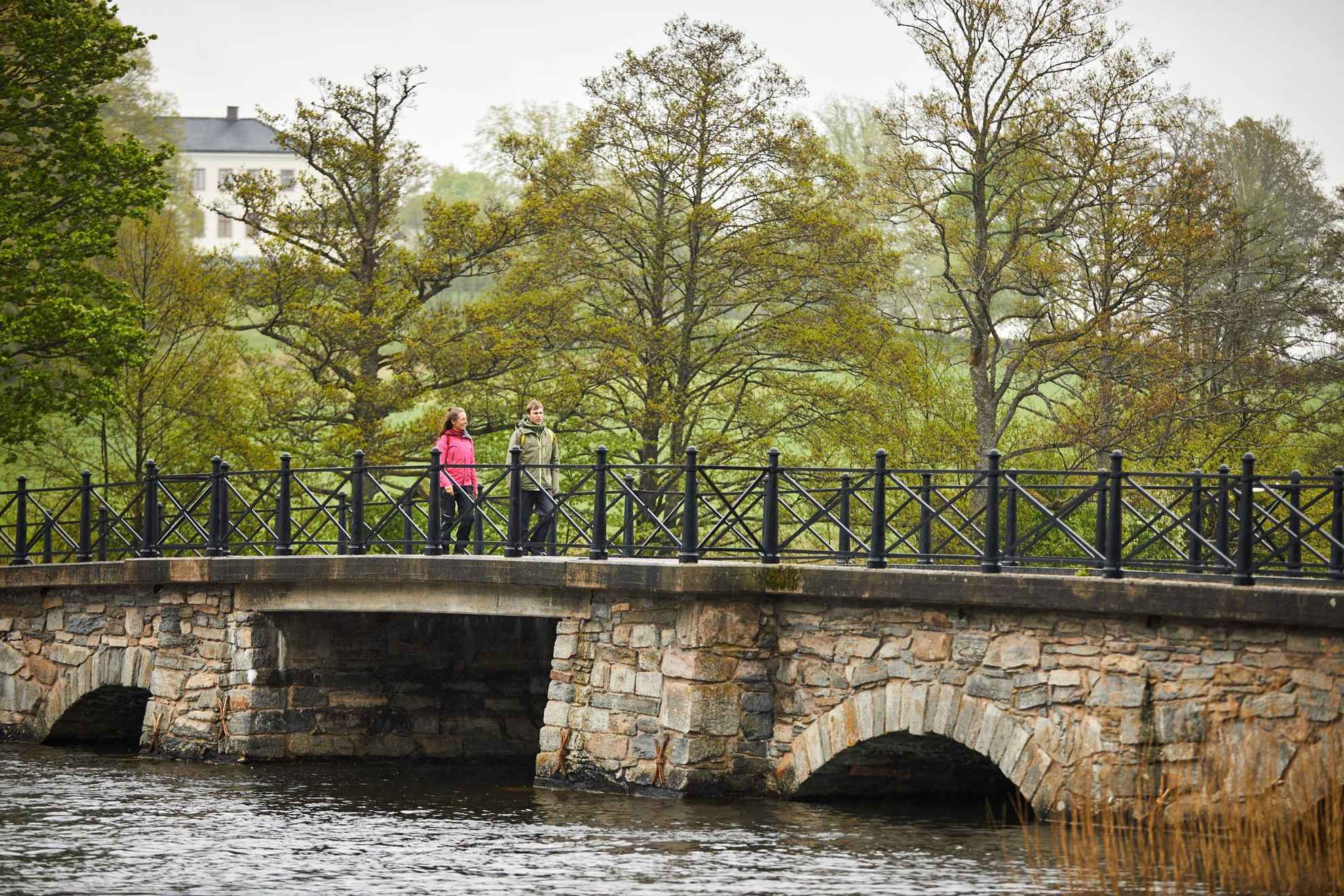 A man and a woman walking on an old stone bridge over a river. There are trees on the riverside and a white house in the background.