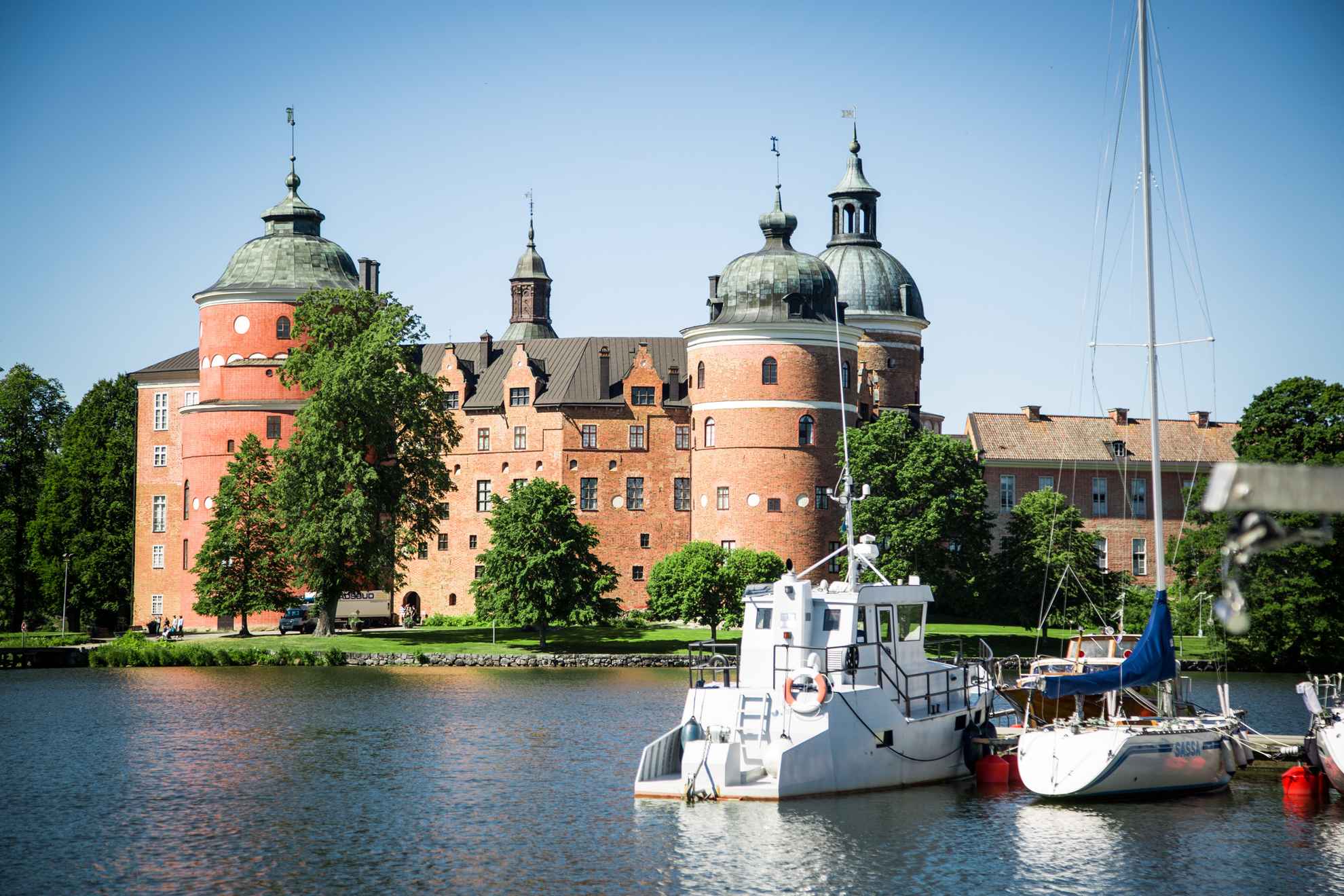 A castle surrounded by greenery is located at the other side of a lake. The sun is shining and boats are docked by the pier.