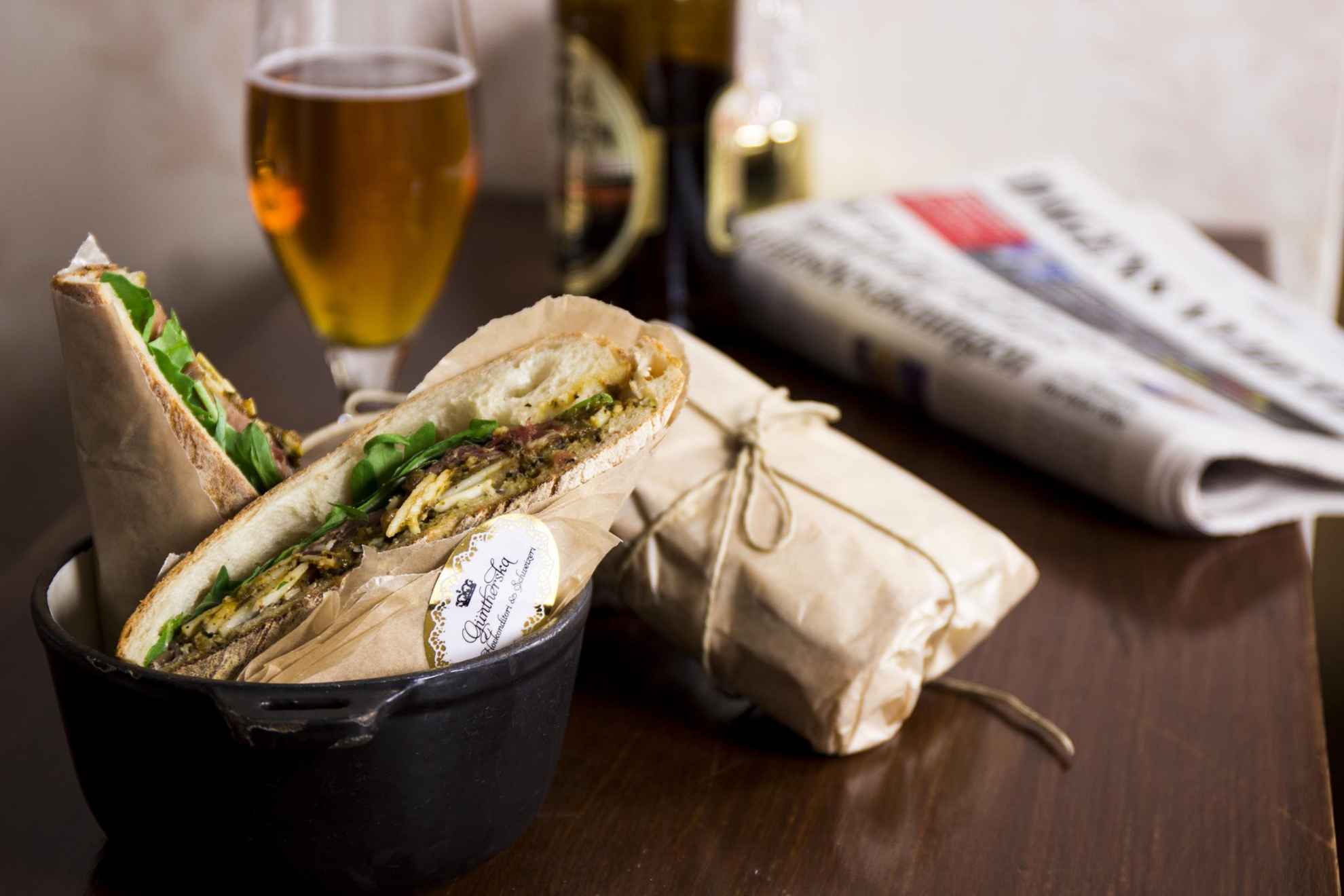 Close-up on a table with a sandwich, a beer and a newspaper.