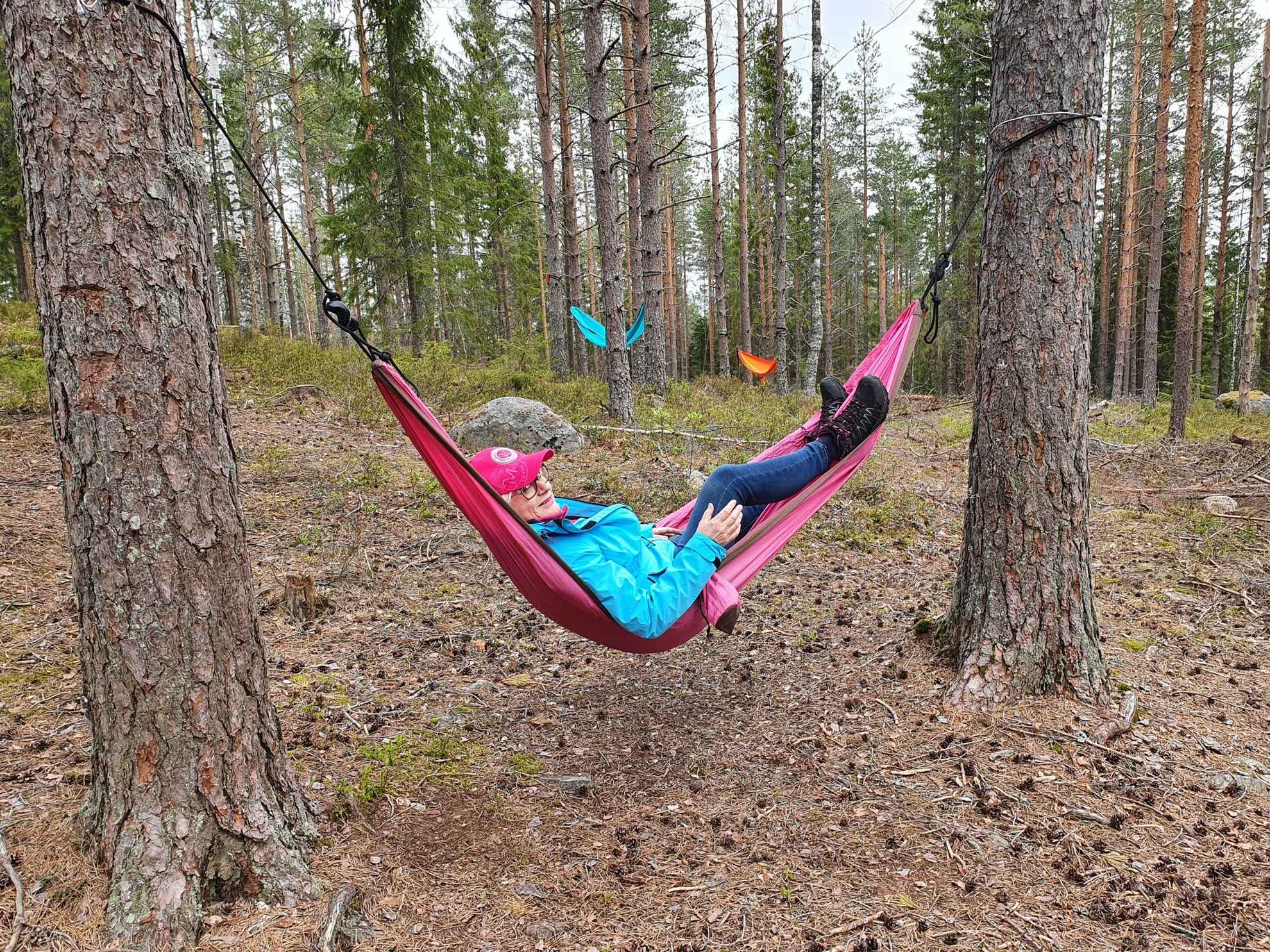 A woman relaxes in a pink hammock between two pines. There is a blue and an orange hammock between the trees in the background.
