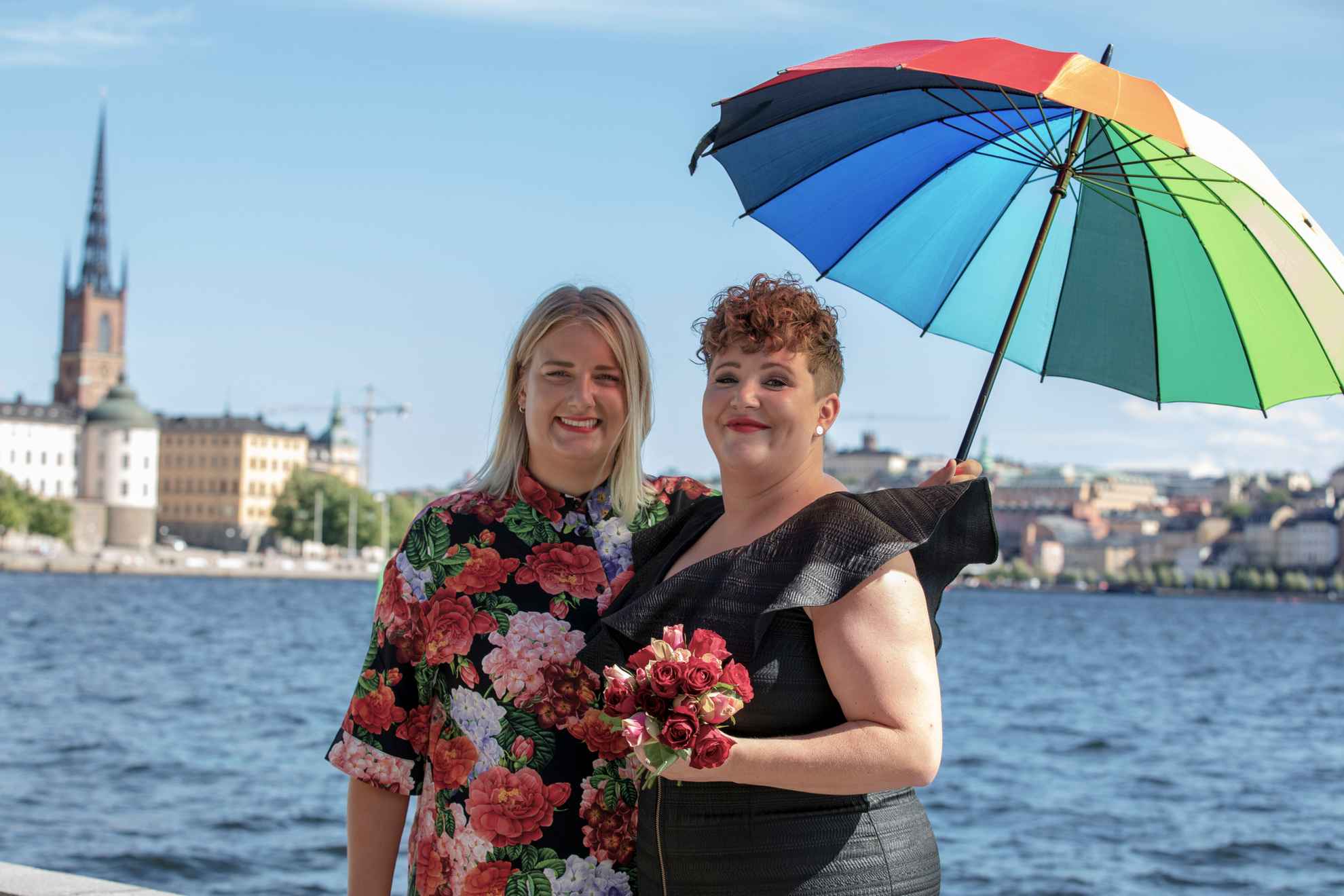 Two women, one with a black dress and one with a flower patterned shirt, are holding an umbrella whit the colors of the rainbow. They are standing by the water with a view of Stockholm city in the background.