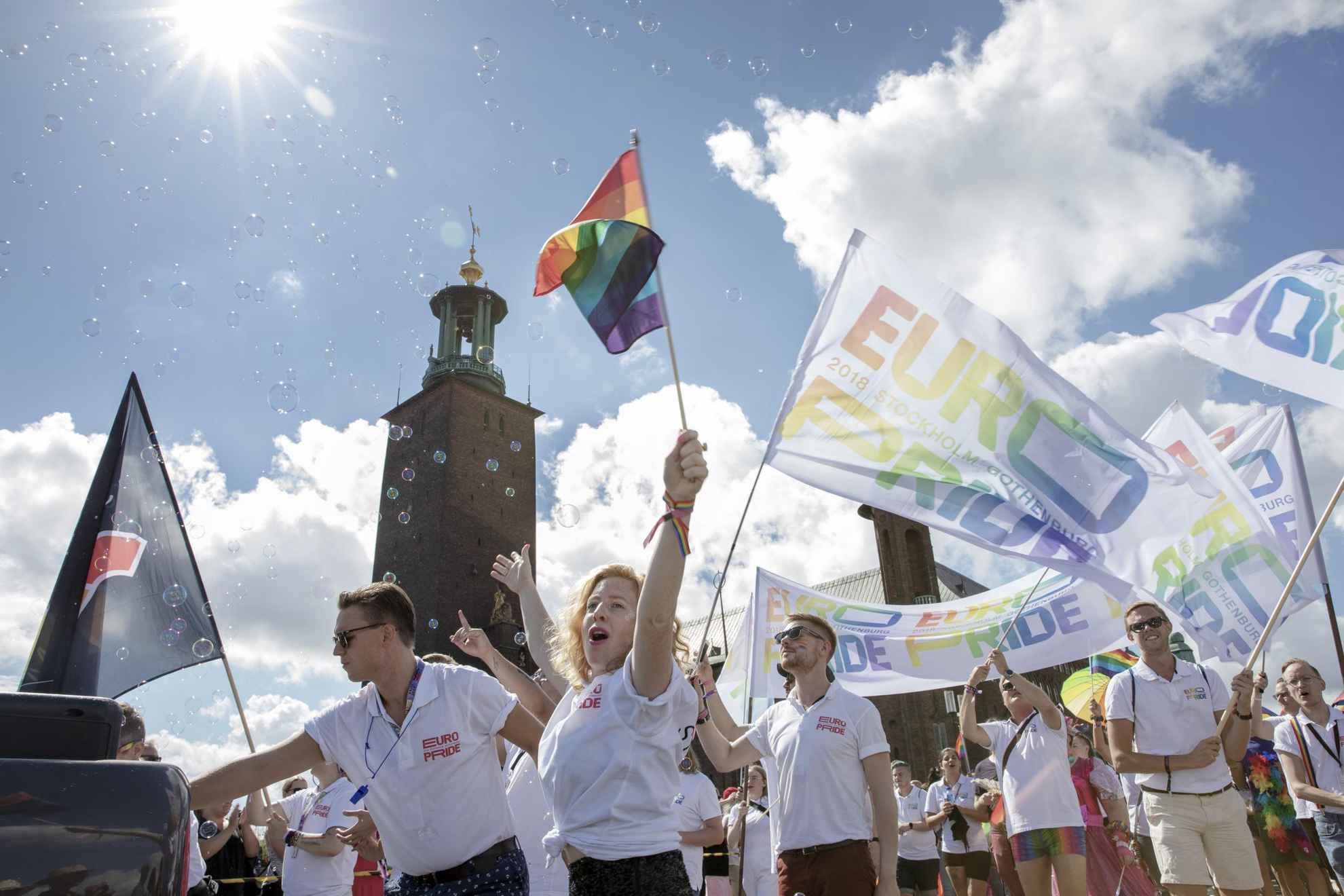 Several people walking in a Pride parade. The have rainbow flags and flags with the text Euro Pride. Stockholm city hall in the background.