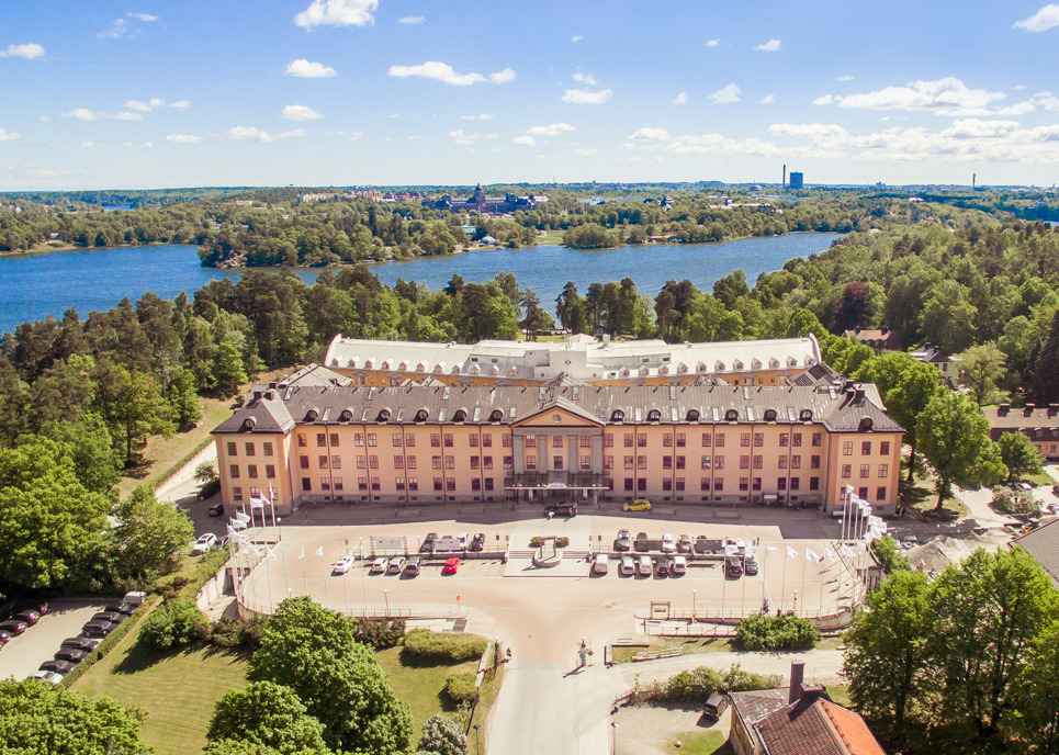 Aerial view of Hagastrand with Brunnsviken shimmering in the background and parked cars visible in the foreground on a sunny day.