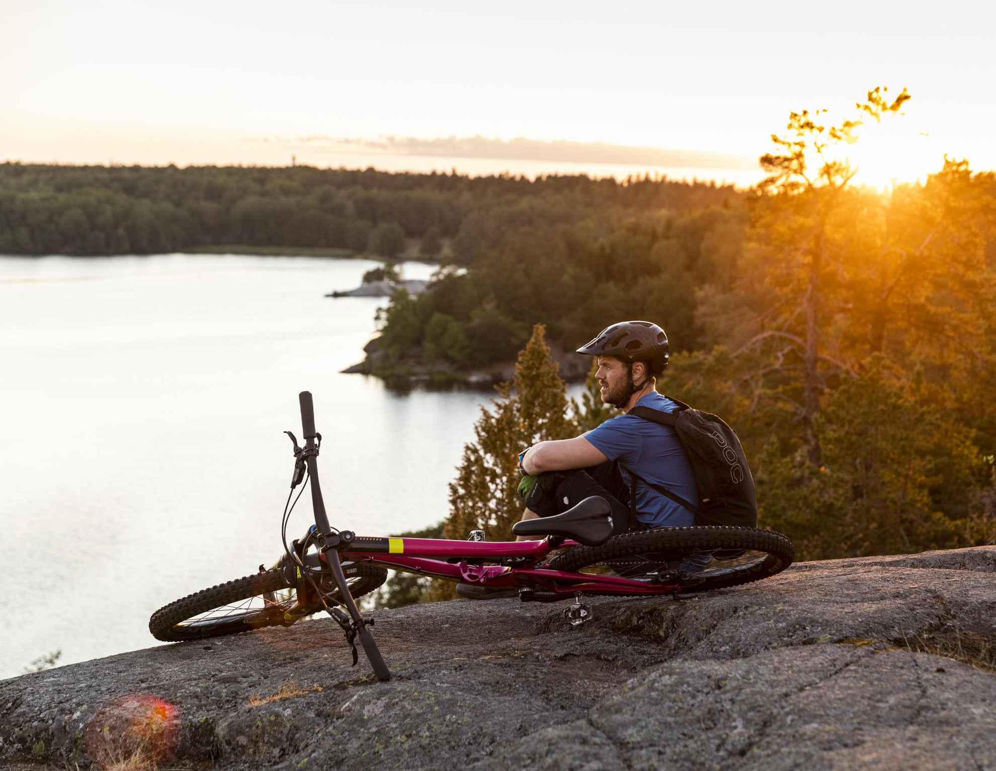 A man is relaxing on a cliff with his bike lying next to him. The man is overlooking a lake.