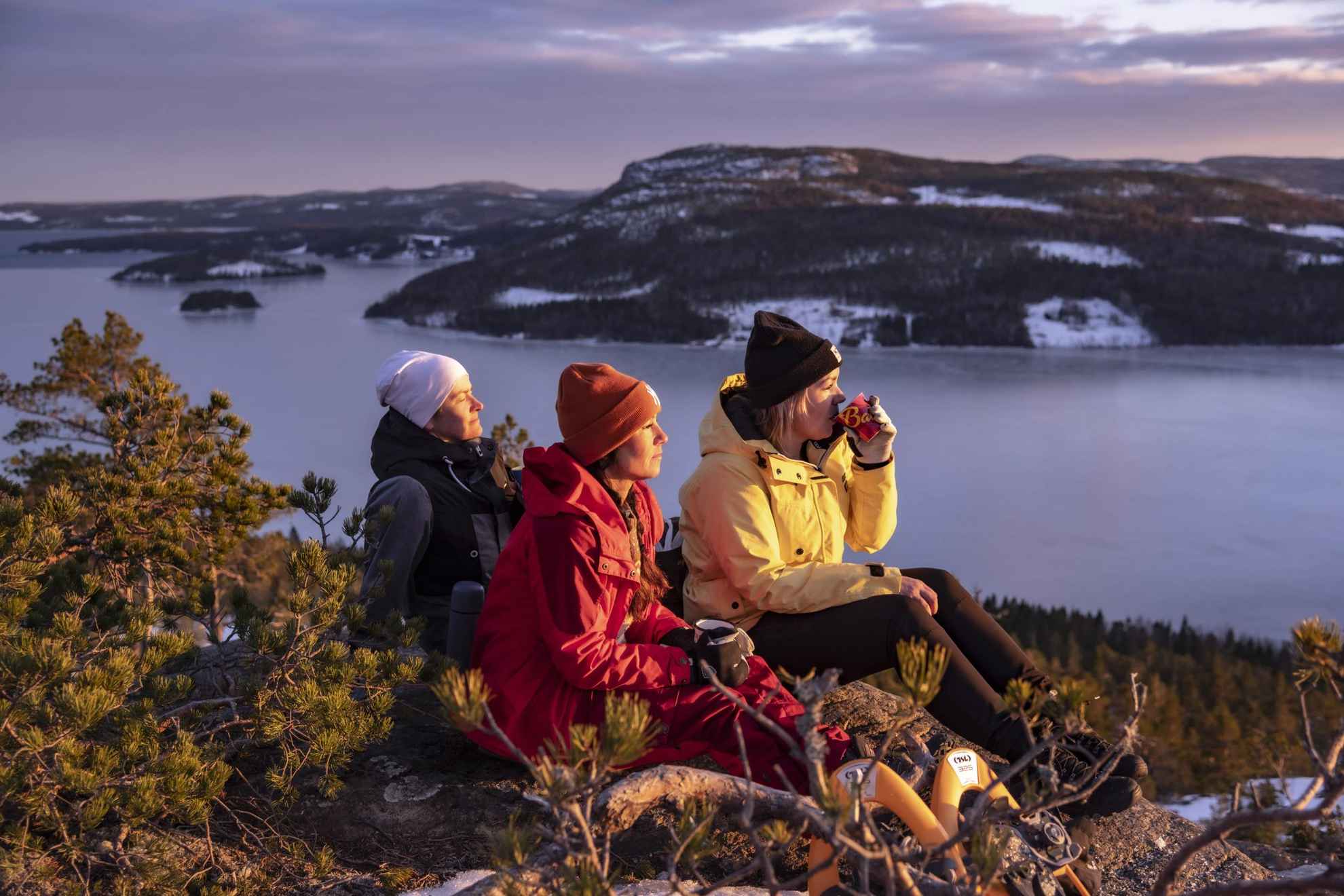 Three people are enjoying the sun with a cup of coffee on top of a hill during winter.