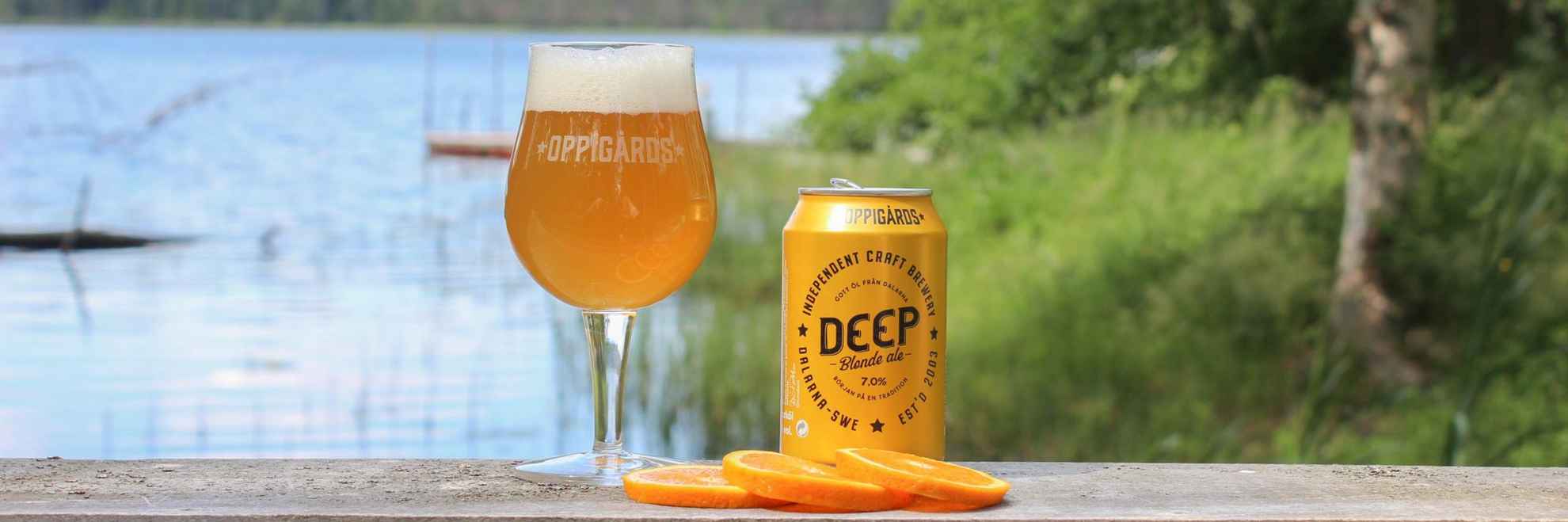 A glass of beer, a can of beer called DEEP and some slices of orange on a table with water and greenery in the background.