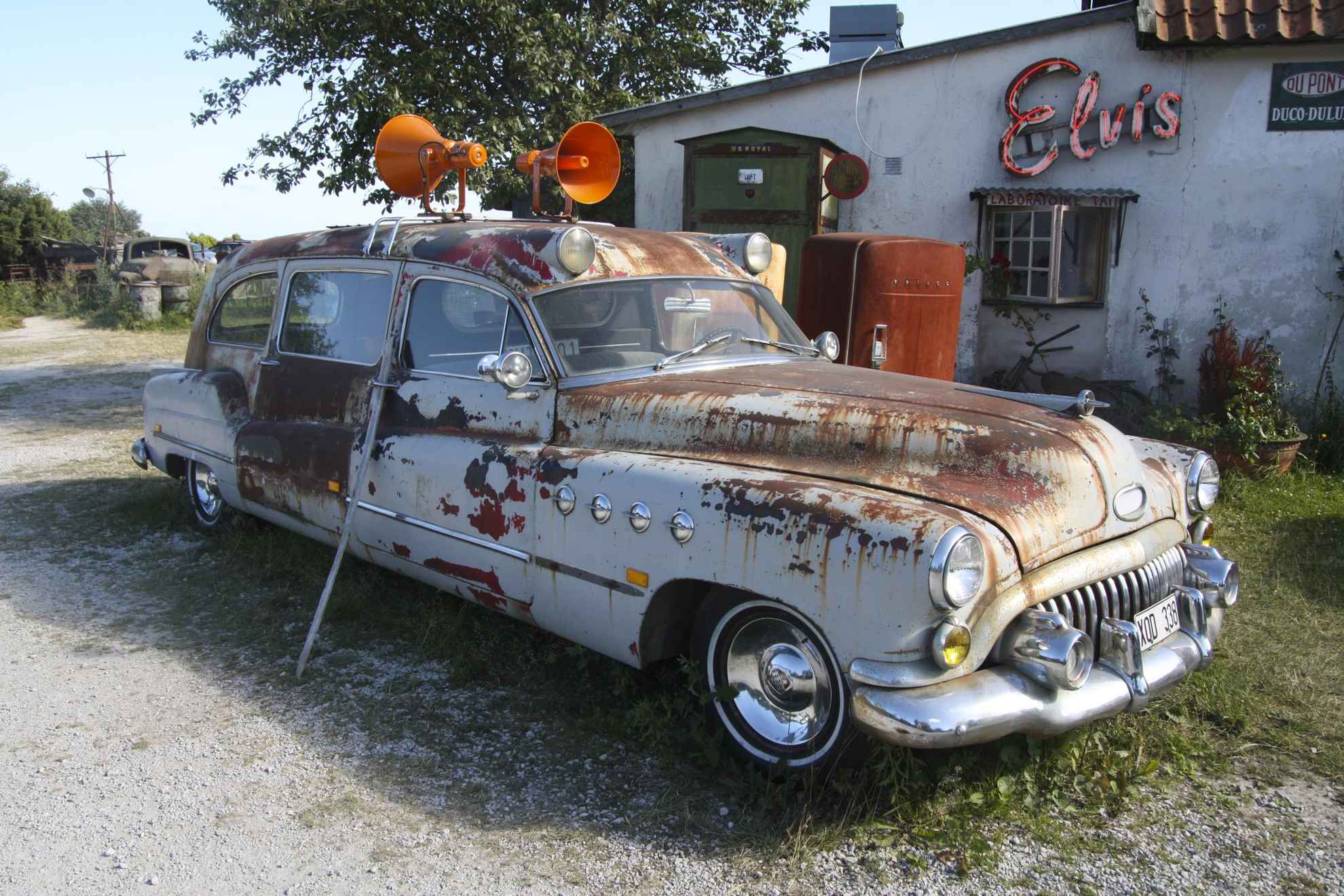 An old rusty car with sirens on the roof in front of an old gasoline station.