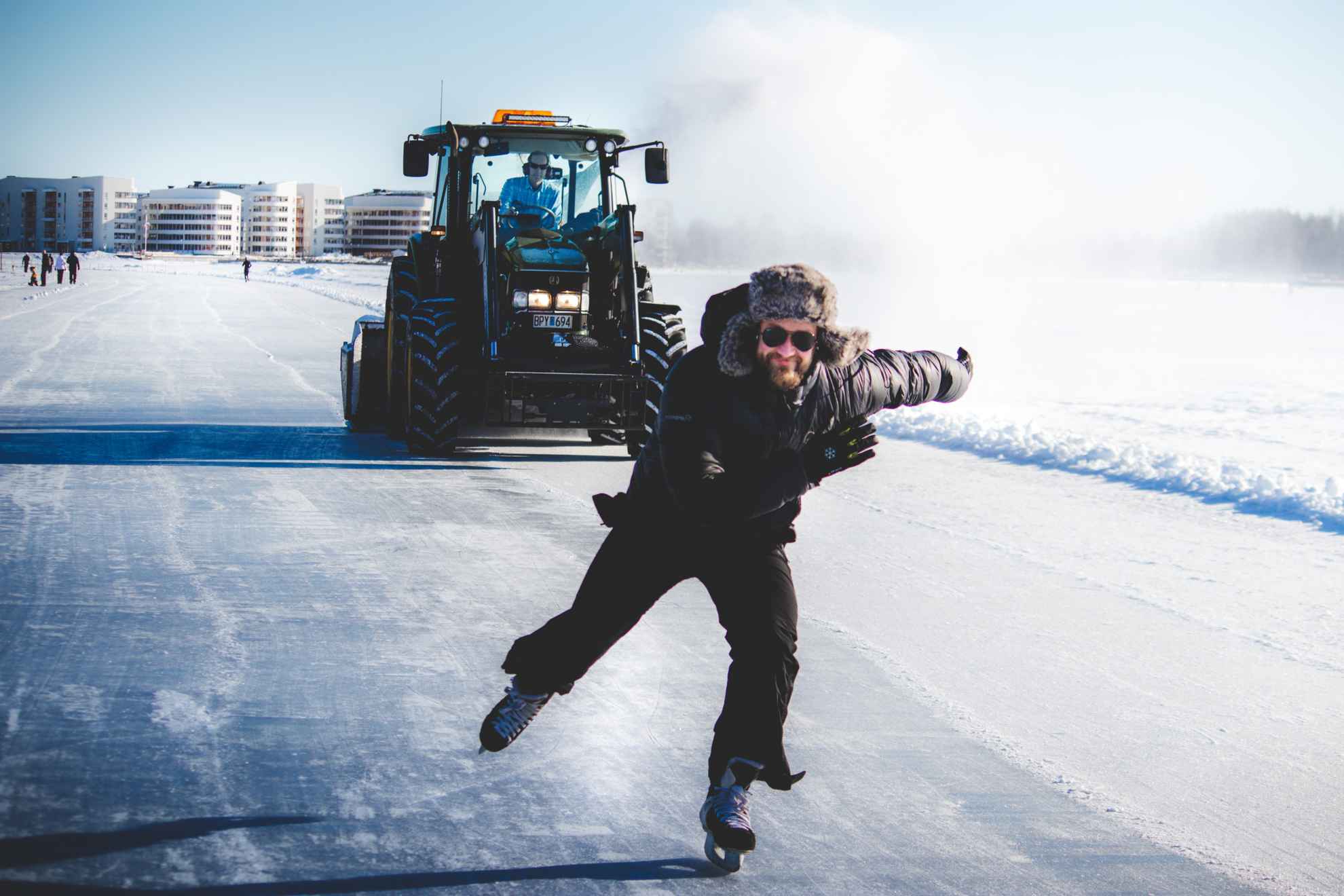 Man dressed in black skating on ice with a tractor behing him