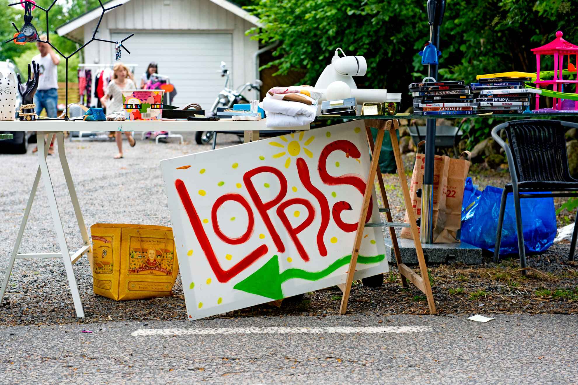 A sign is leaned against a table with an assortment of used items. A house is in the background.