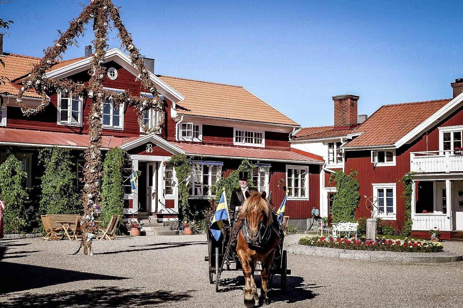 A large red wooden house with white edges during the summer. In front of the house are a midsummer pole and a horse with carriage with two Swedish flags.
