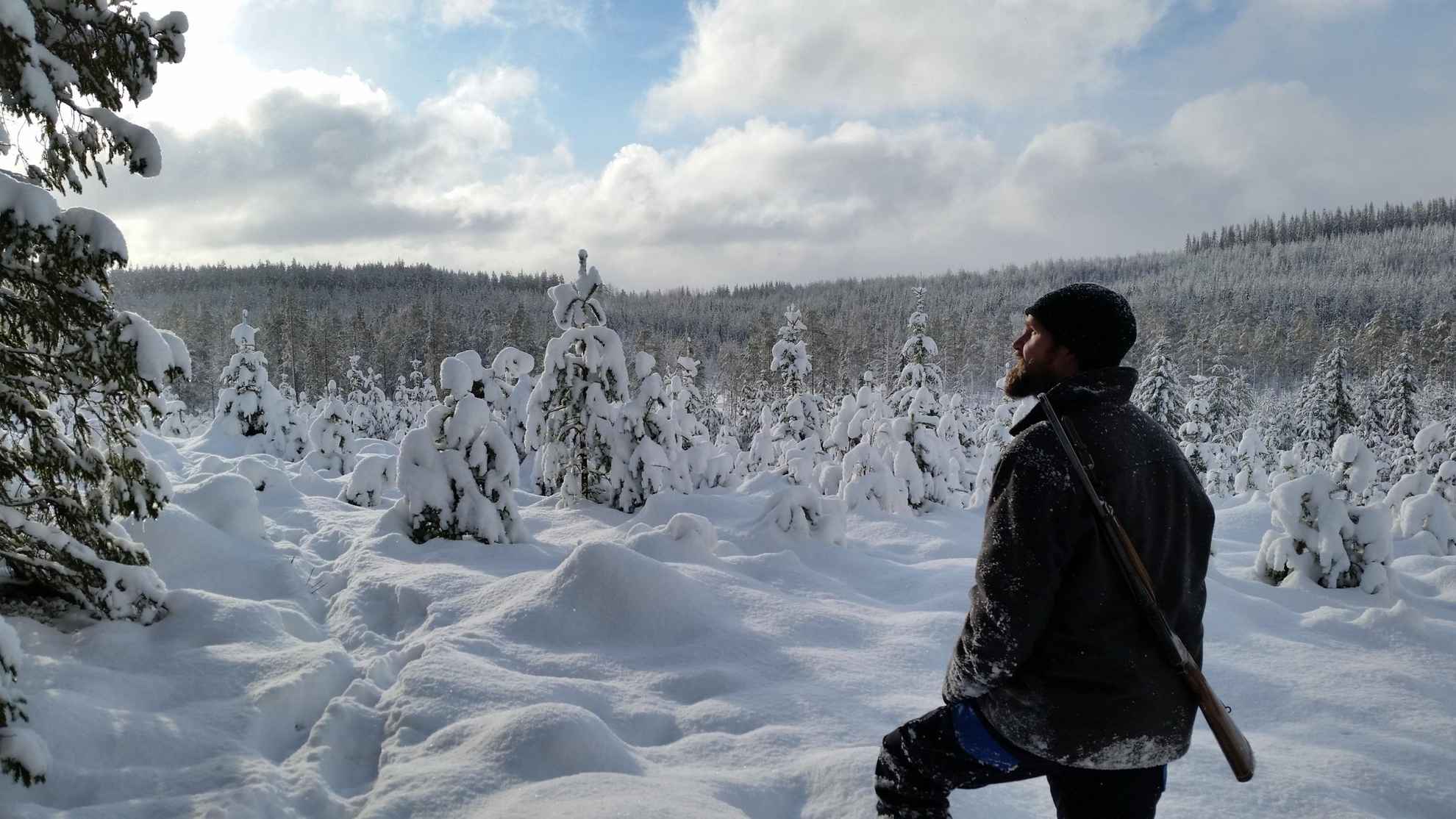 A man is standing in a snow-covered forest with a hunting rifle hanging on his back.