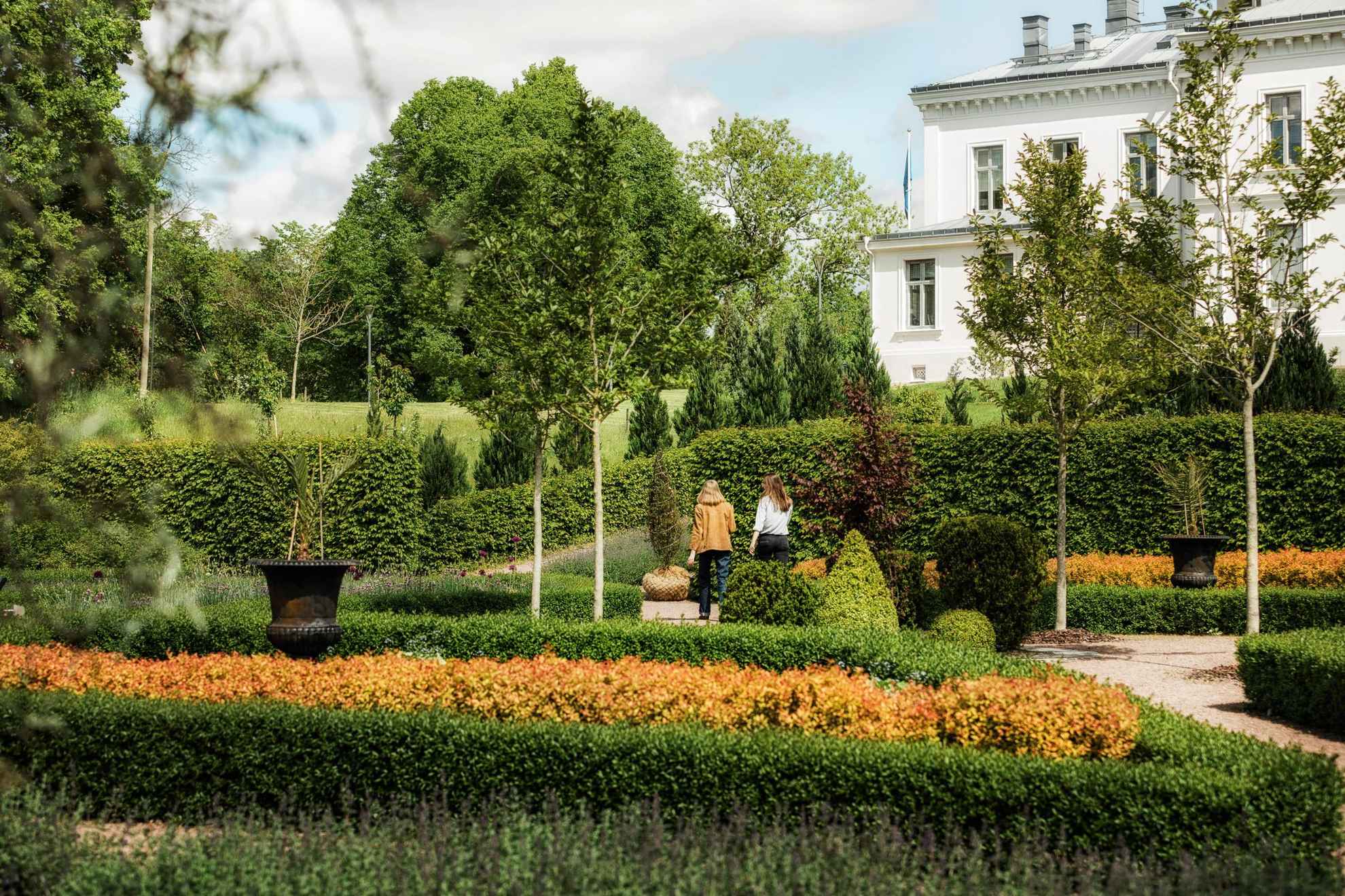 A beautiful arrange garden with orange plants between green bushes. A white 19th century manor house to the right and two women walking towards the house.