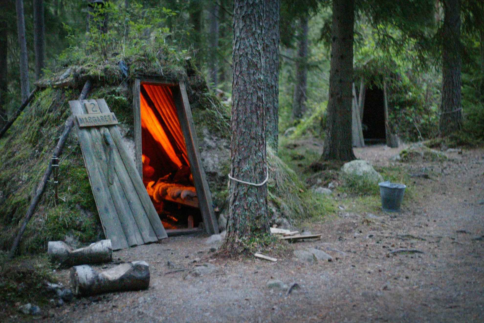 Two forest huts covered with moss located in a forest. In one of the huts a fireplace is burning.